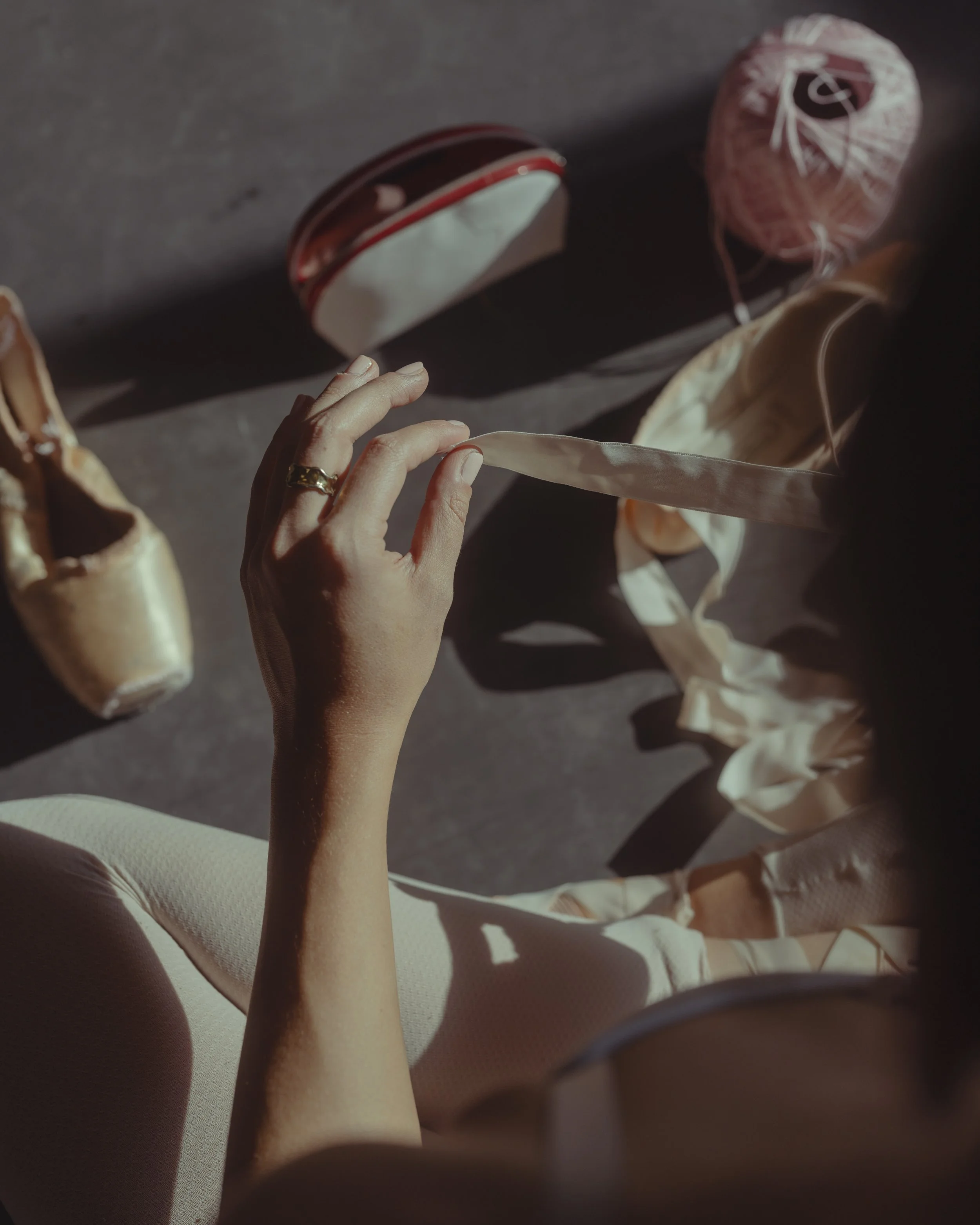 A hand with a gold ring holds a satin ribbon, with sewing supplies such as a pin cushion, ball of yarn, and fabric scattered on a dark surface in natural light.