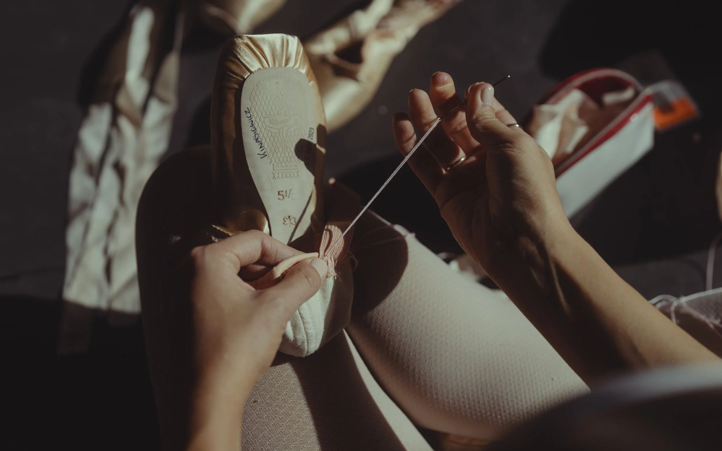 A person sewing the sole of a beige shoe with a needle and white thread, with a box of sewing supplies nearby.