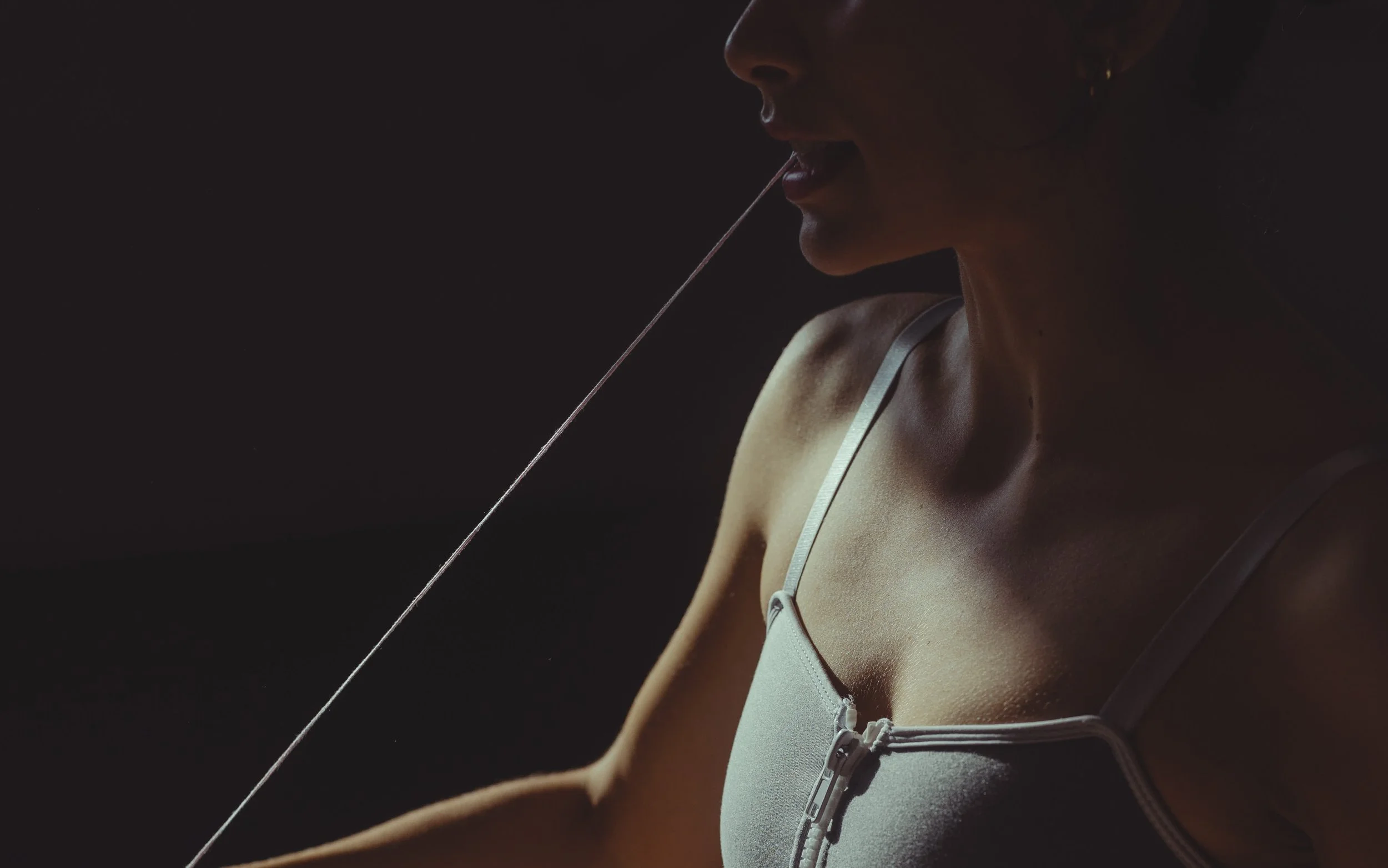 Close-up of a woman with a draped tank top, touching her lips with her finger, with a string or thread near her mouth, against a dark background.