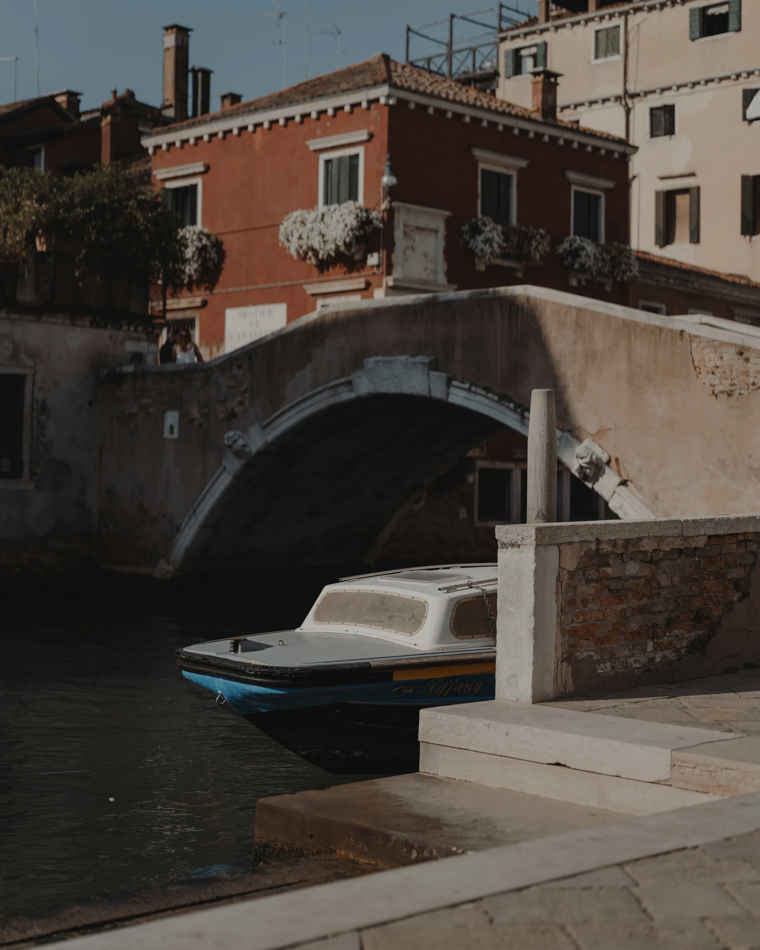A small boat docked along a canal with a bridge and colorful buildings in the background, in Venice, Italy.