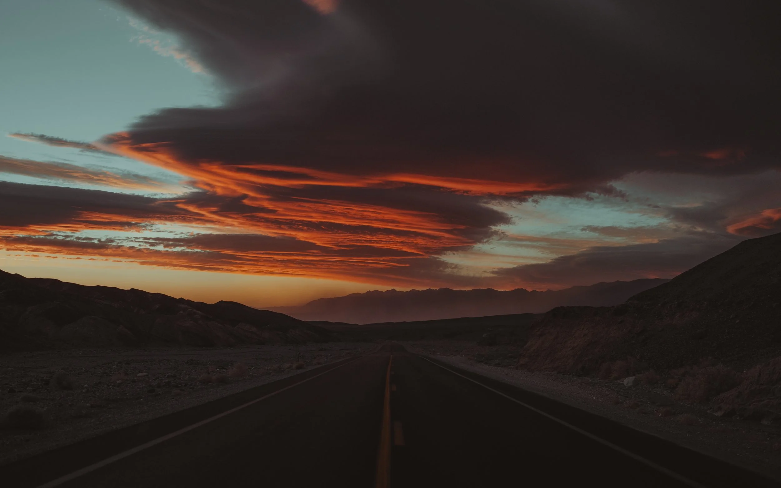 A straight road extending into the distance through a mountainous landscape during sunset, with dramatic orange, red, and dark clouds in the sky.