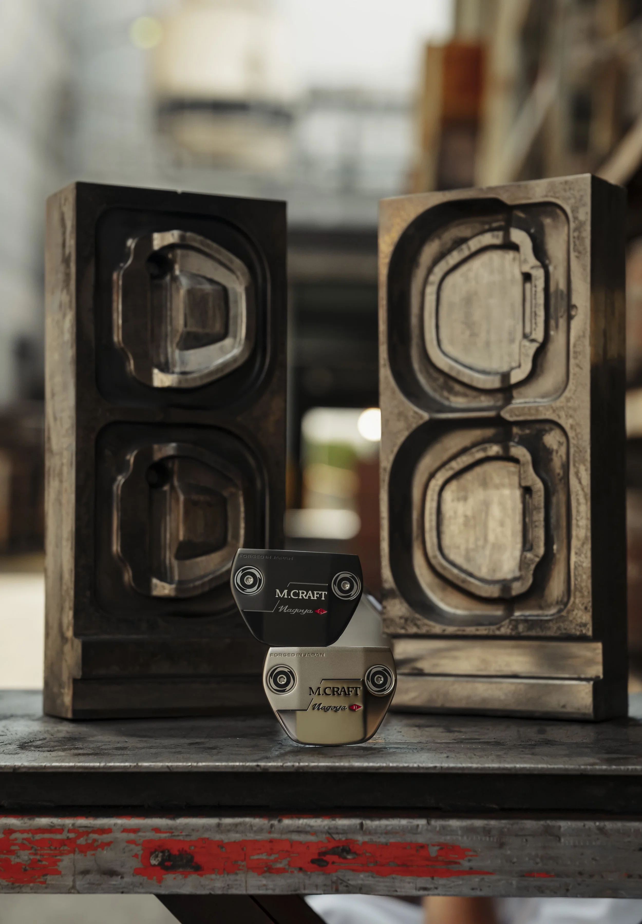 Two metal objects with the label »M. CRAFT« and »Magoya« are placed on a worn wooden surface. They are positioned in front of a large, industrial-looking, rectangular metal mold with four cavities, which appears used and somewhat rusty. The backgroun