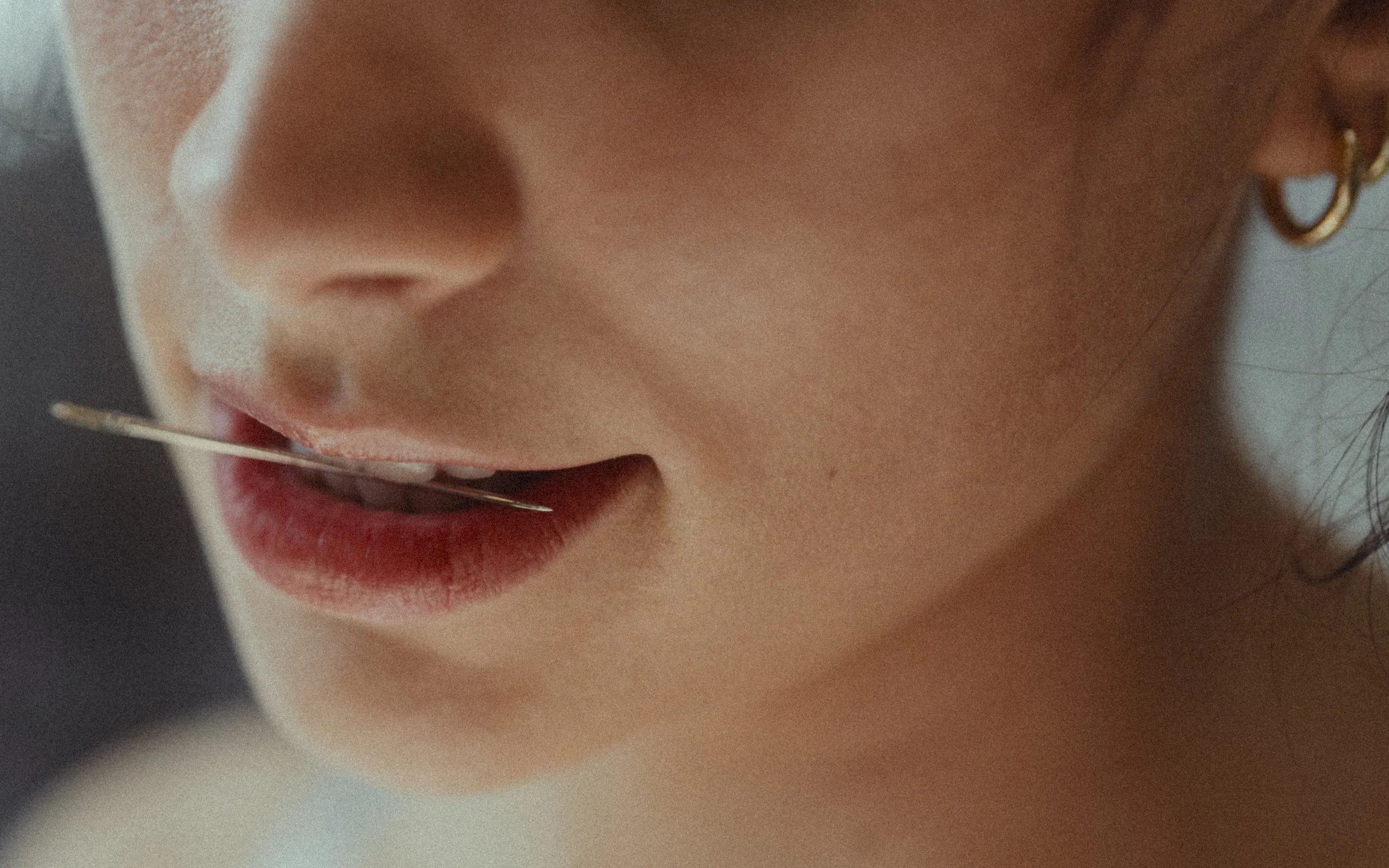 Close-up of a person's face with a pin in their mouth, showing lips, nose, and part of an earring.