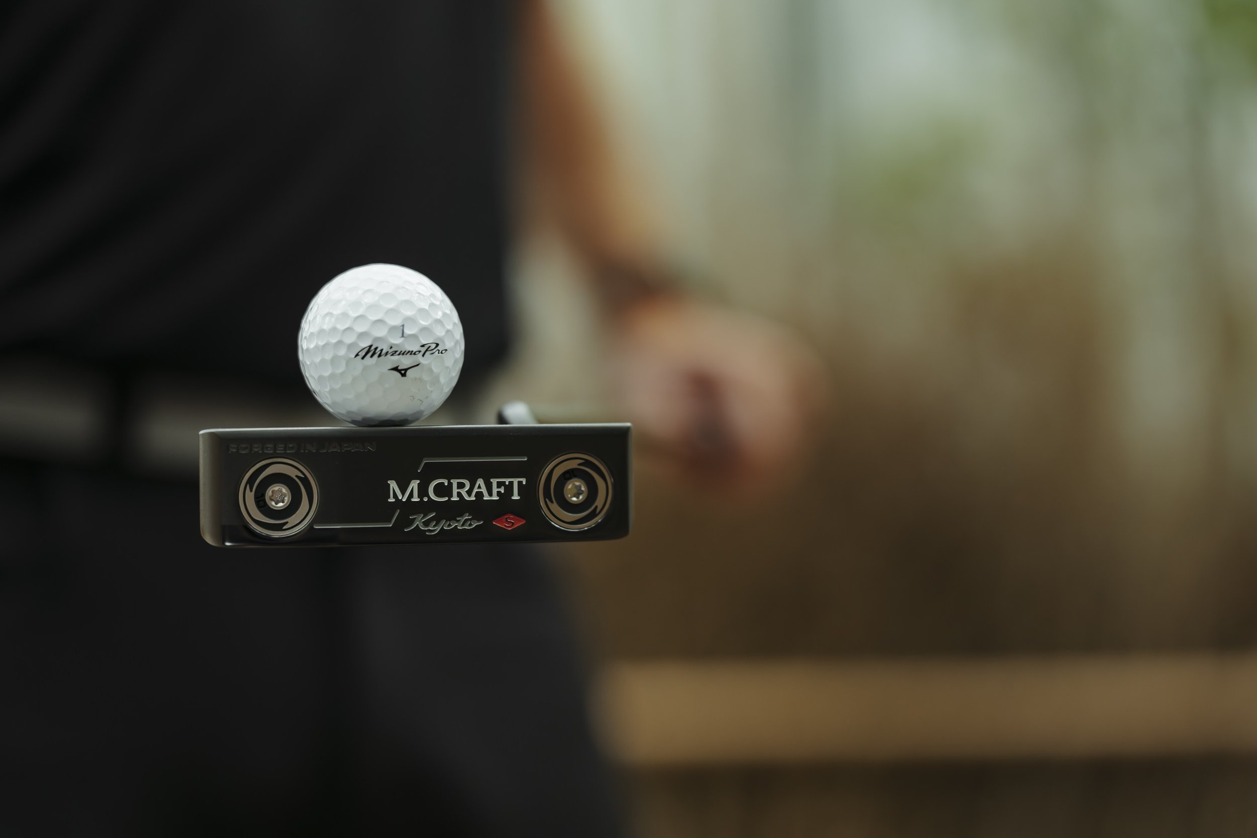 A white golf ball labeled 'Mizuno Pro' resting on a black golf club with 'M. CRAFT Kyoto' written on it, outdoors with a blurred background.