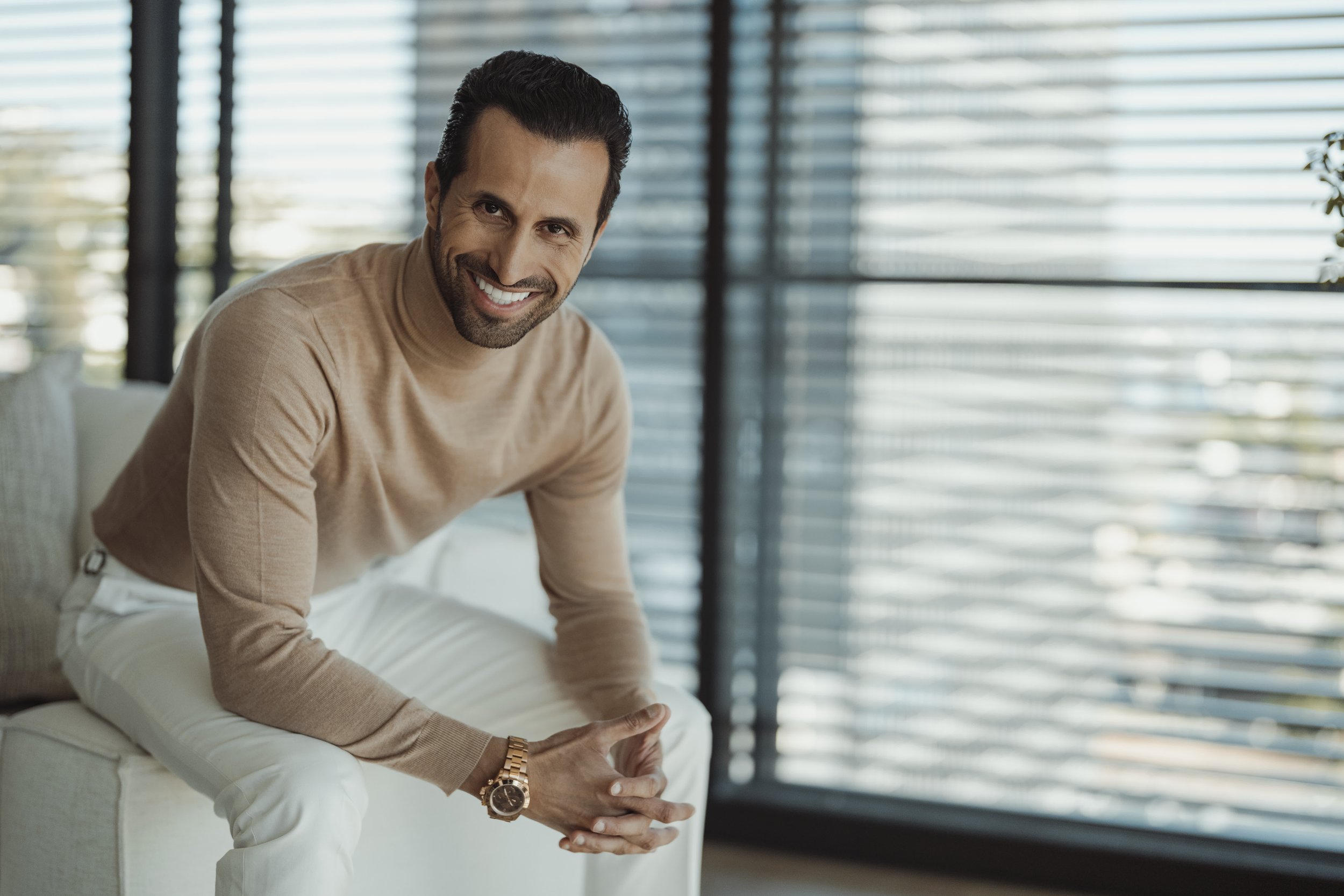 A smiling man with dark hair and a beard, wearing a tan sweater, white pants, and a gold watch, leaning forward on a light-colored sofa inside a room with large windows and horizontal blinds.