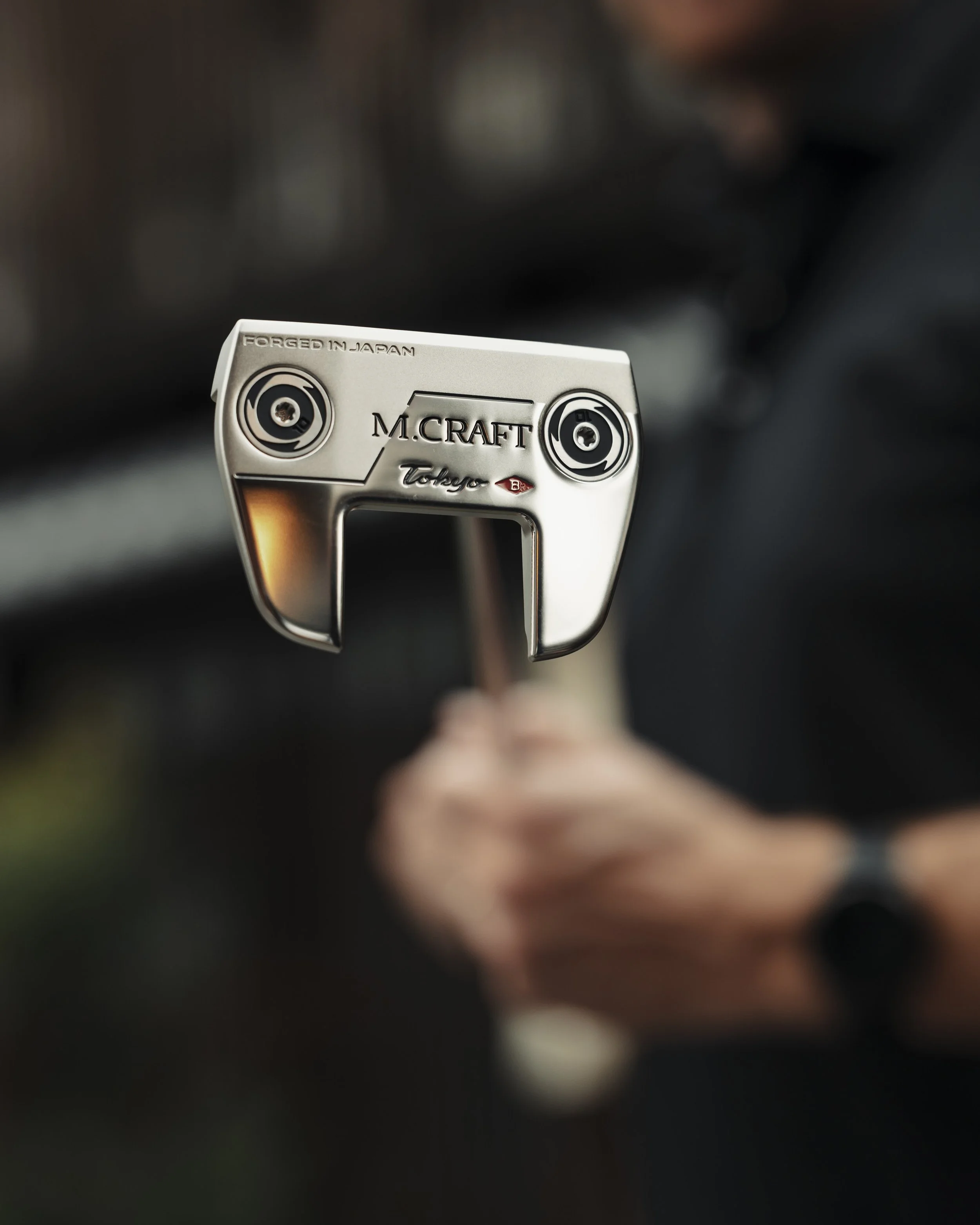 Close-up of a golf putter head engraved with 'M. CRAFT' and 'Tokyo', being held by a blurred person in the background.