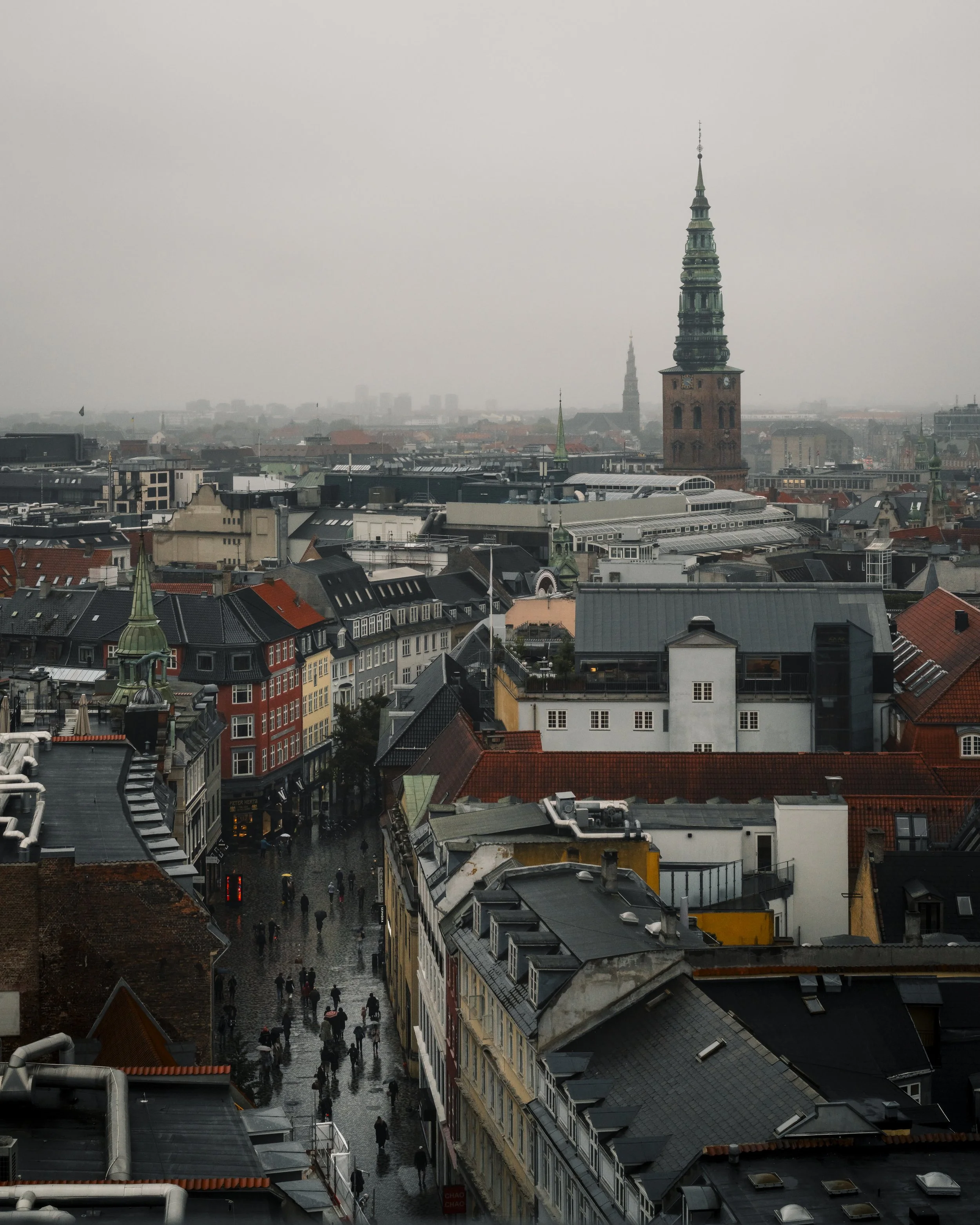 A cityscape on a rainy day with colorful buildings, a tall church steeple, and pedestrians walking with umbrellas.