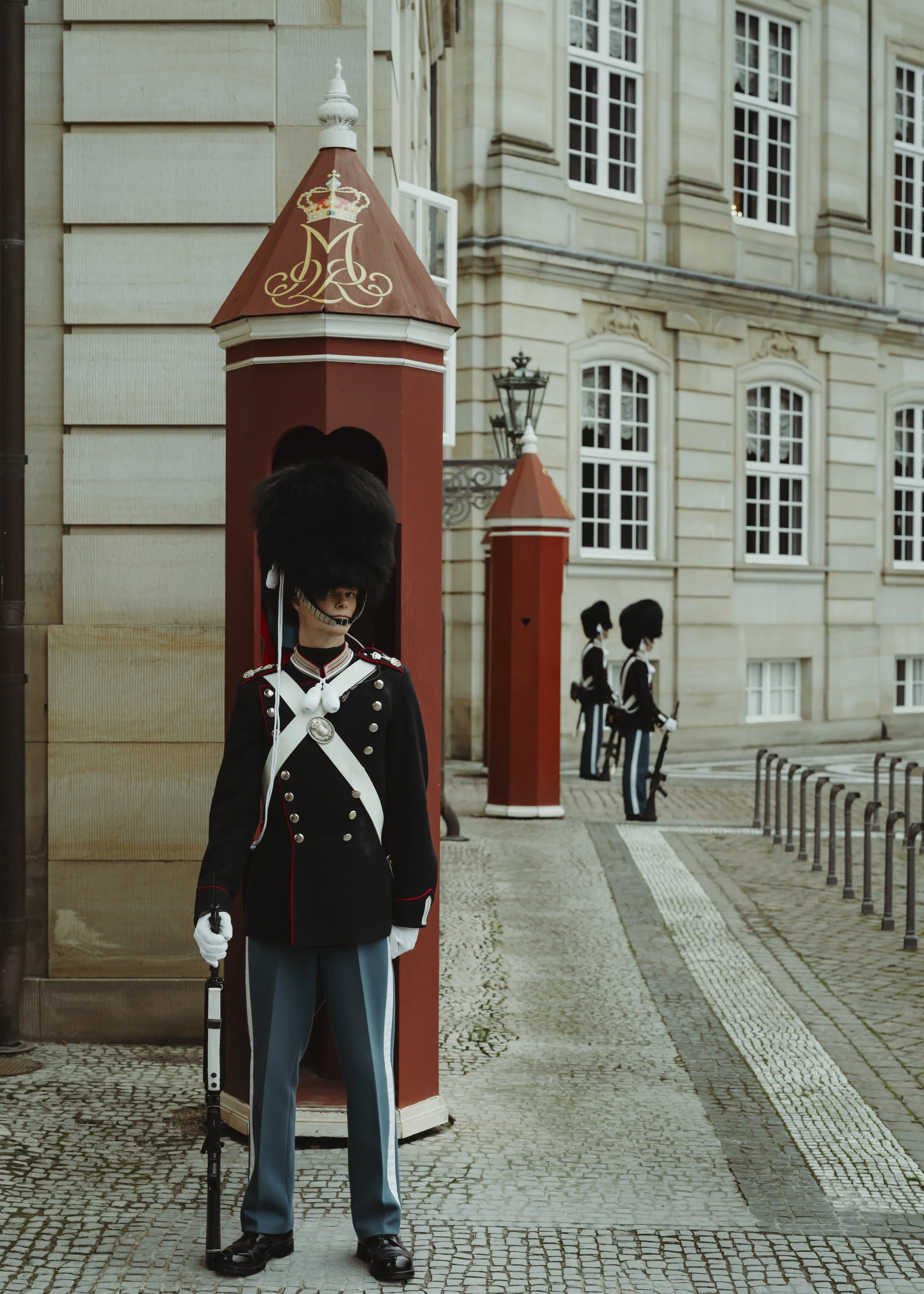 Soldier in traditional uniform with bearskin hat standing guard outside a historical building, with two other guards in similar uniforms in the background.