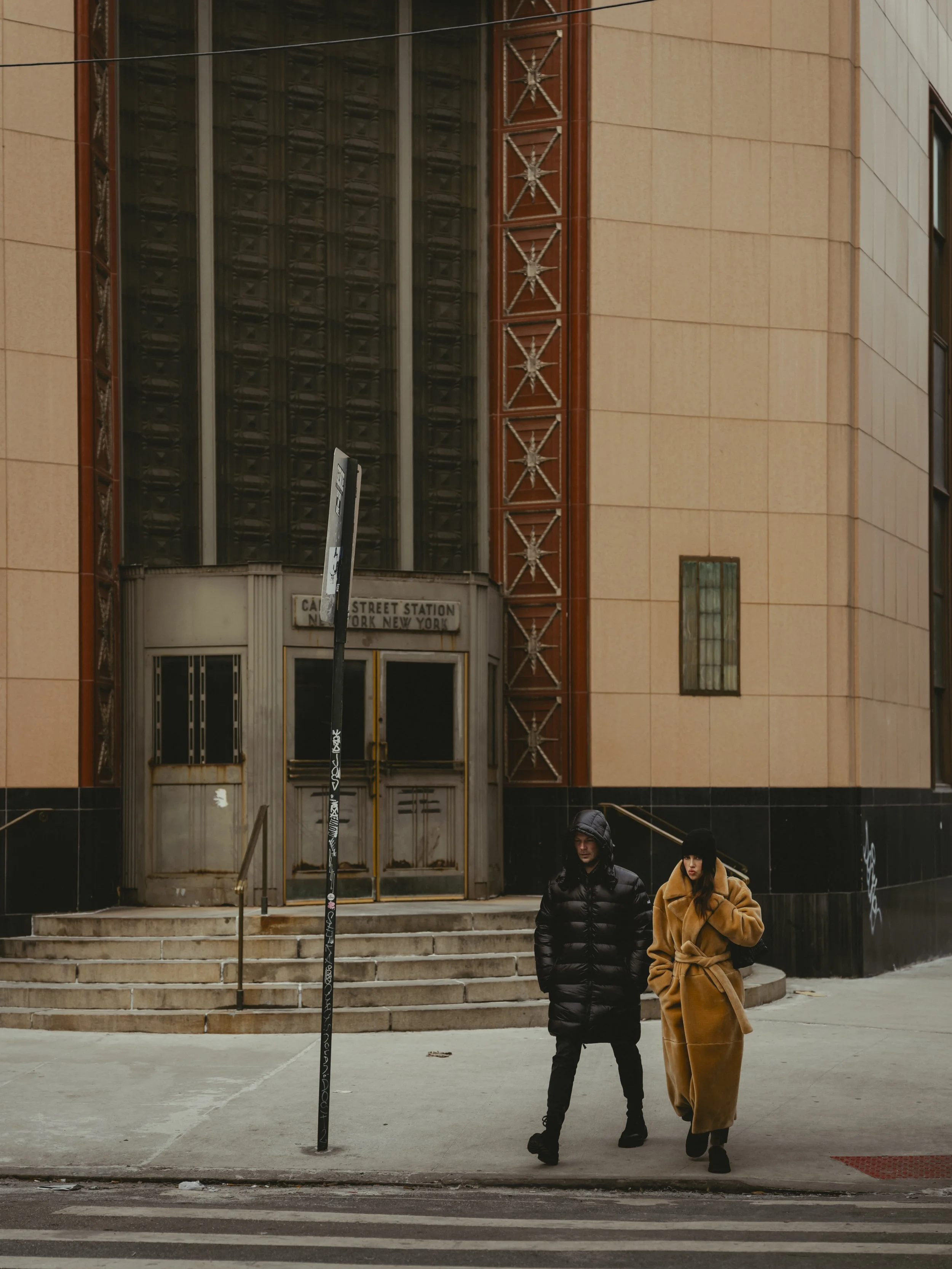 Two people walk on the sidewalk outside an old building with stairs leading to a door, in front of the 125th Street Station in New York City during cold weather, both dressed in warm coats and hats.