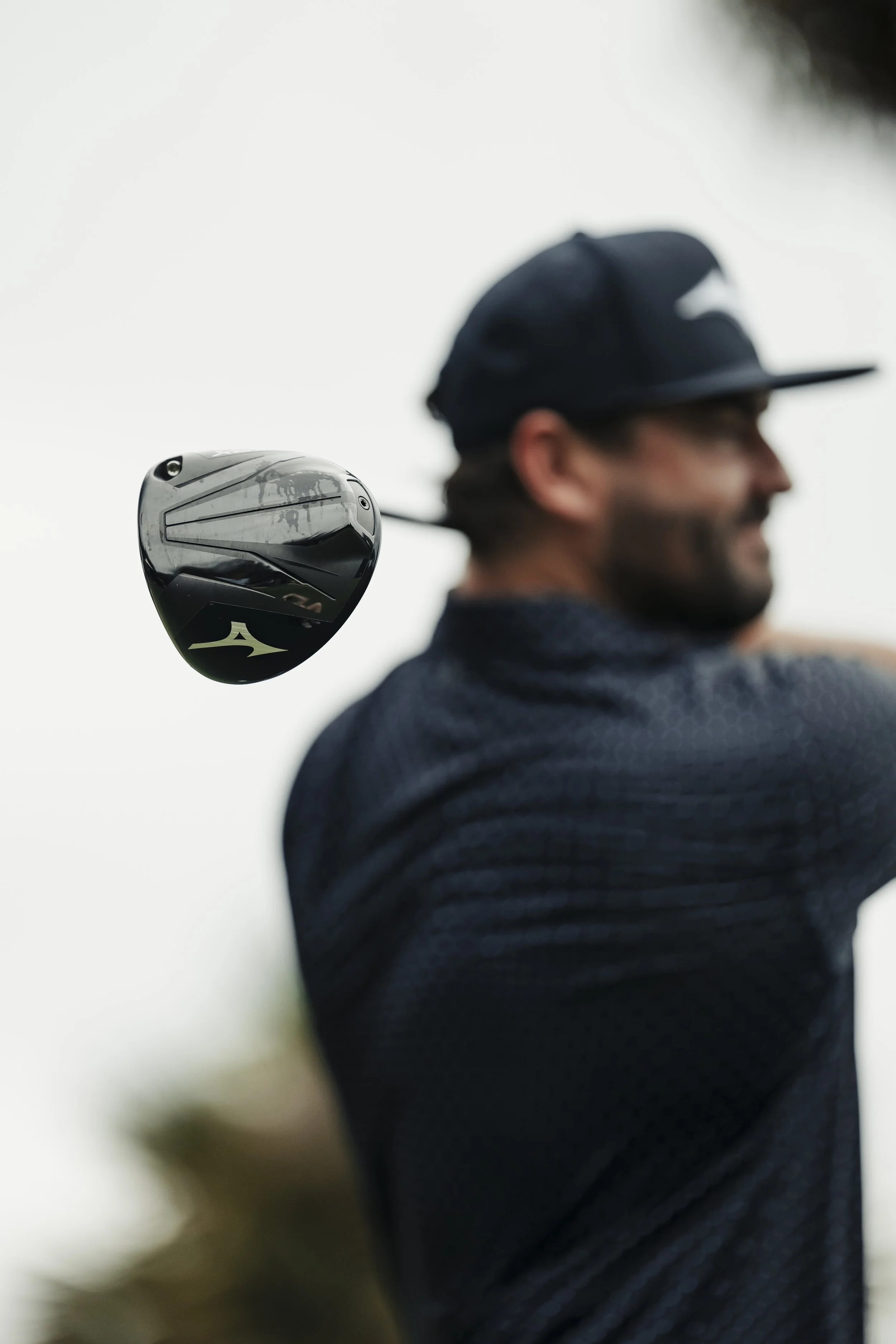 A man in a dark cap and textured shirt is swinging a golf club, with a close-up of the club head in focus behind him.