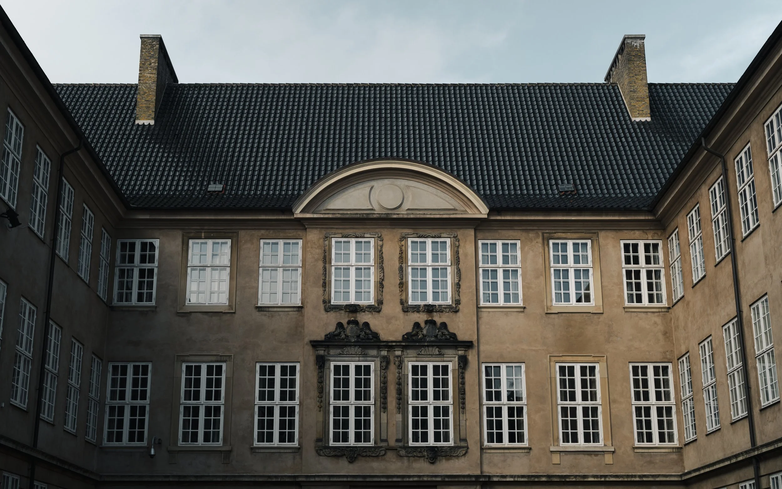 A historic European-style building with multiple windows, a decorative feature above the central windows, a steep black tiled roof, and brick chimneys.