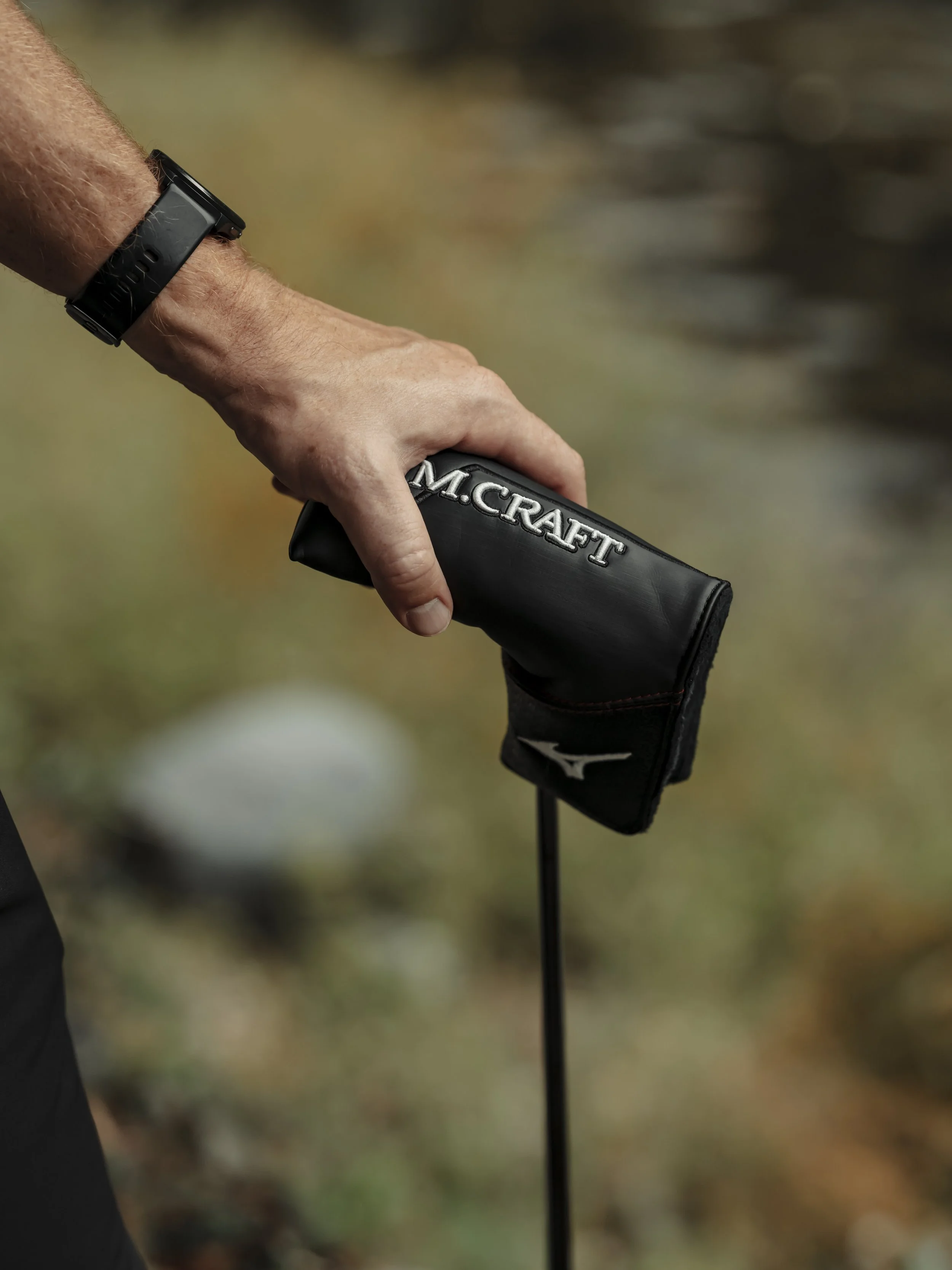 A close-up of a person's hand holding a golf club with a black headcover that has 'M.CRAFT' embroidered on it, set outdoors on a golf course.