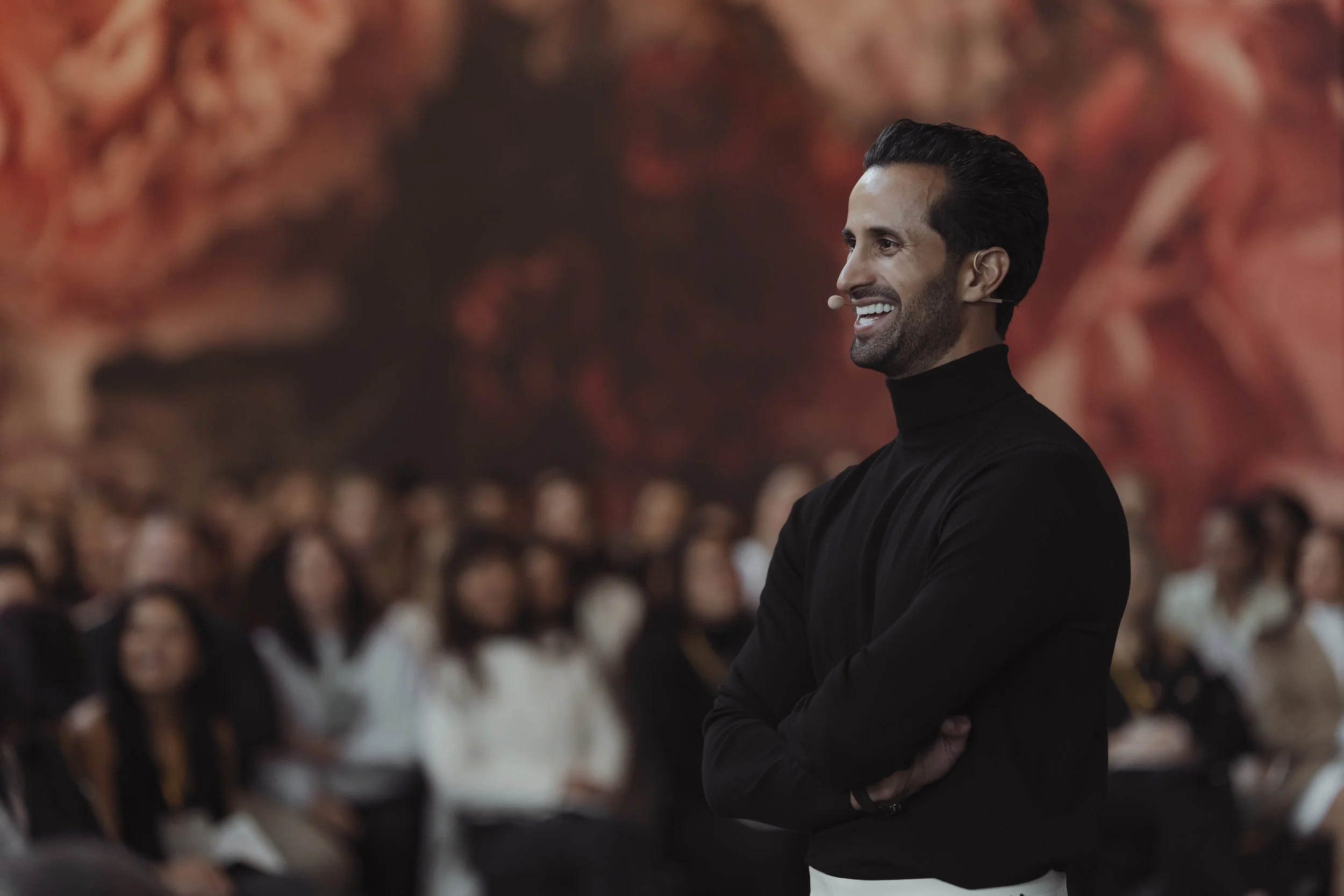 A man with dark hair and a beard, wearing a black turtleneck, smiling and speaking at a conference or presentation with an audience seated behind him.