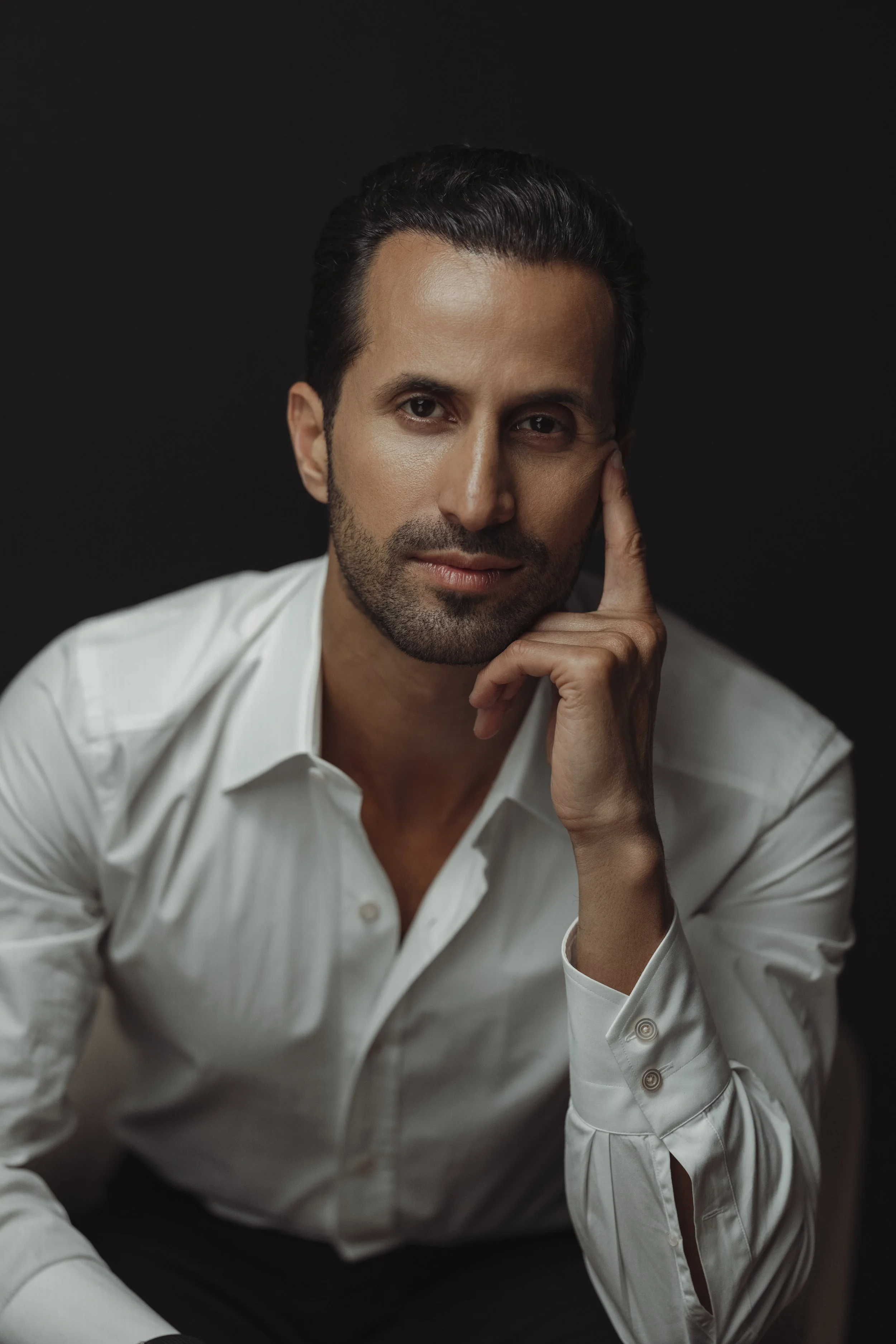 A man with dark hair, a neatly groomed beard, and wearing a white shirt, sitting against a dark background. He rests his chin on his hand, looking directly at the camera with a slight, confident smile.