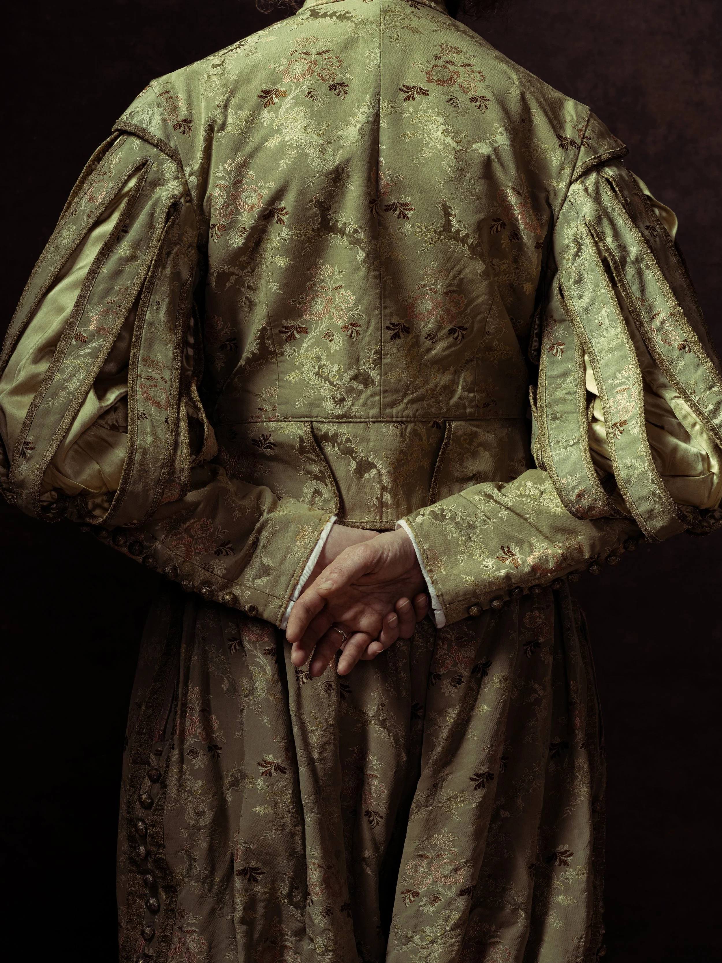 Back view of a person wearing an ornate, gold, floral-patterned garment with long, puffed sleeves, standing against a dark background.
