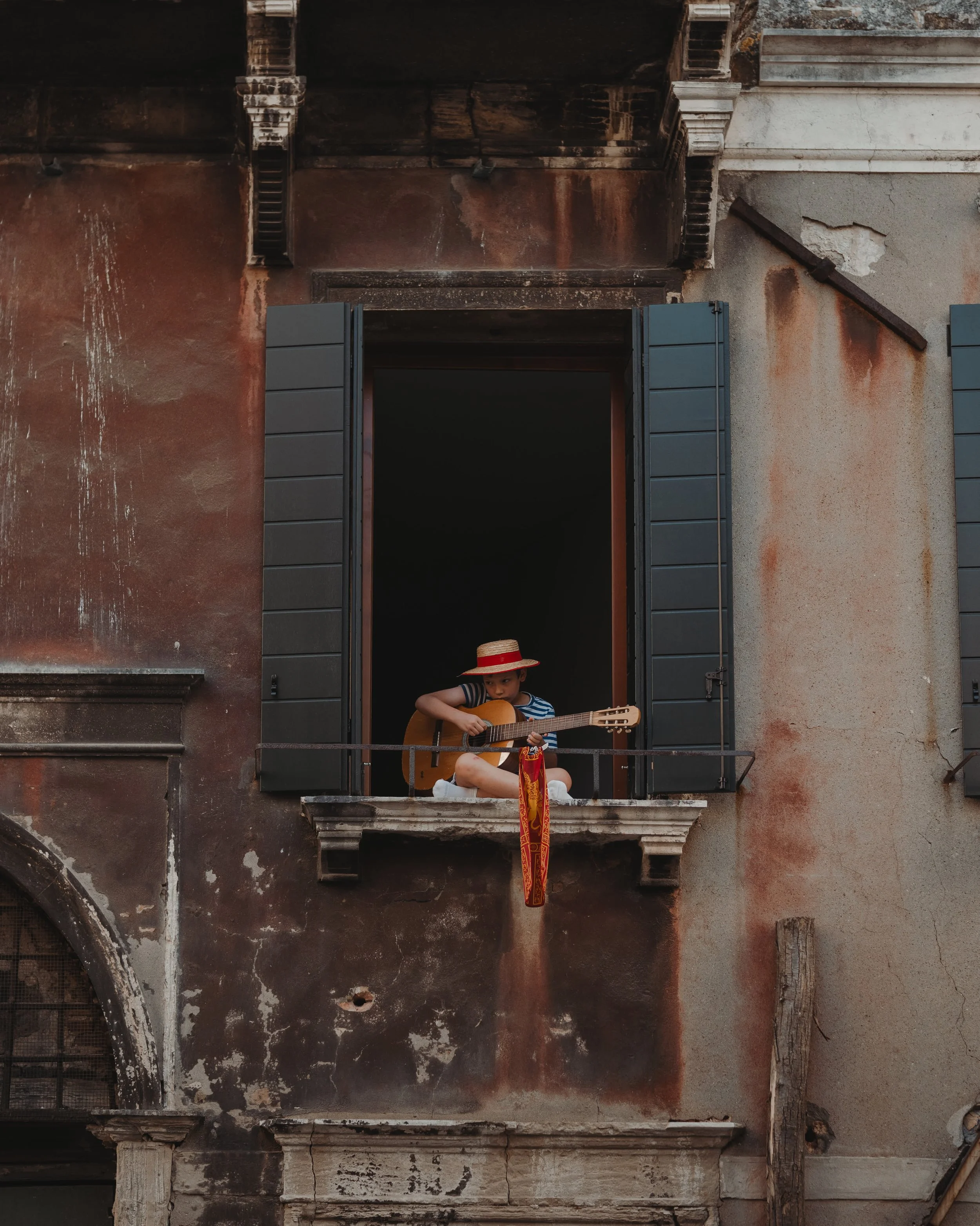 A young boy wearing a straw hat, striped shirt, and shorts sitting on a windowsill playing a guitar with a colorful cloth hanging from it. The building has aged, weathered walls with open shutters.