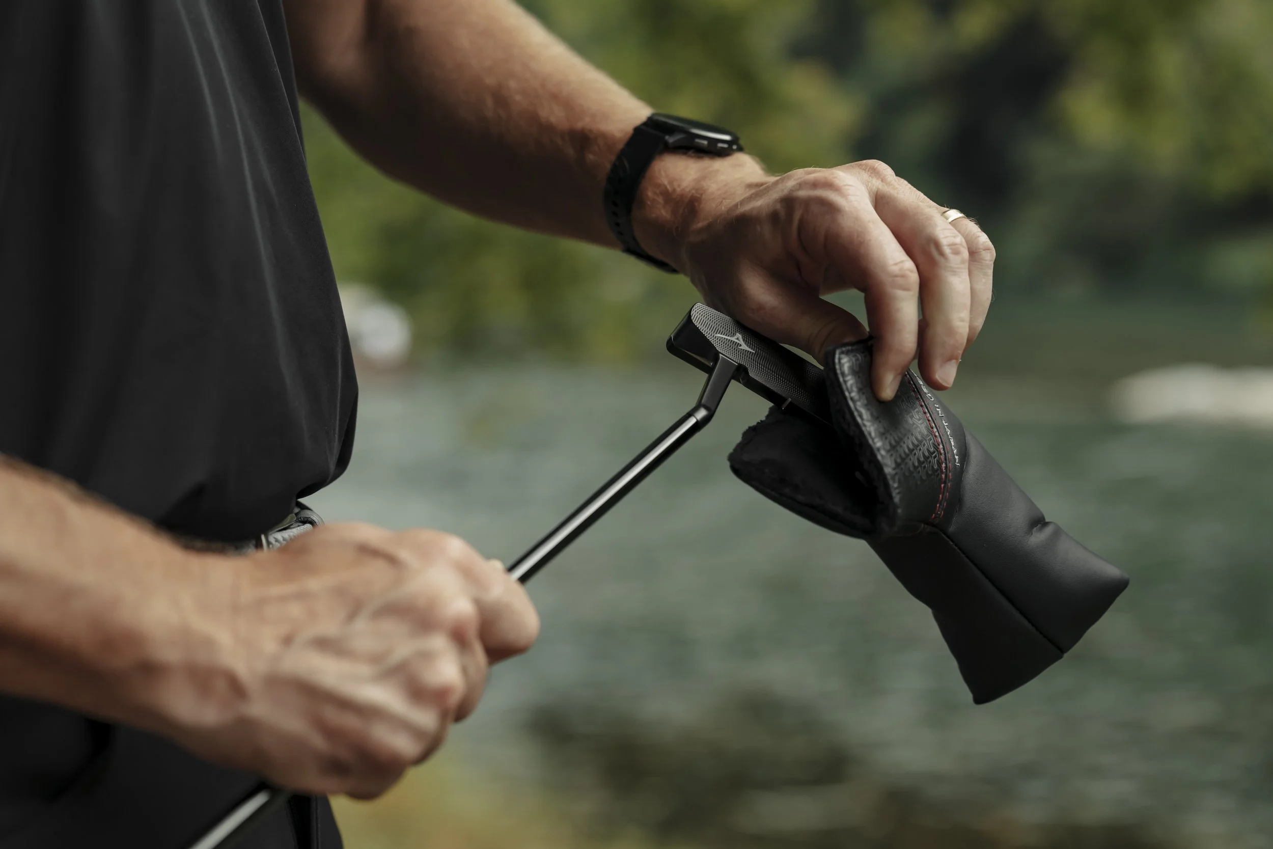 Close-up of a person adjusting a golf club headcover outdoors, with trees and water in the blurred background.