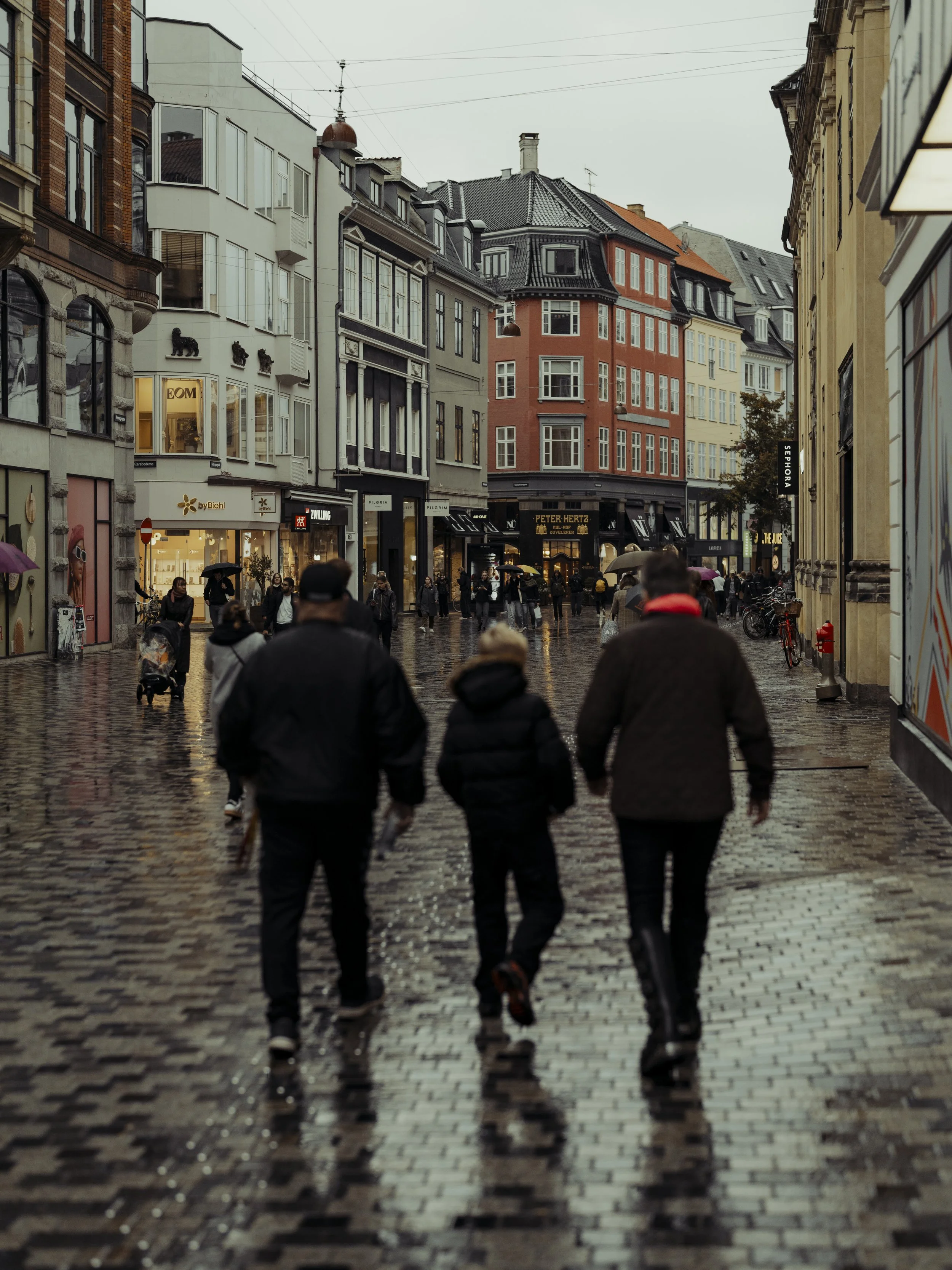 People walking on a wet city street with colorful buildings and storefronts on a rainy day.