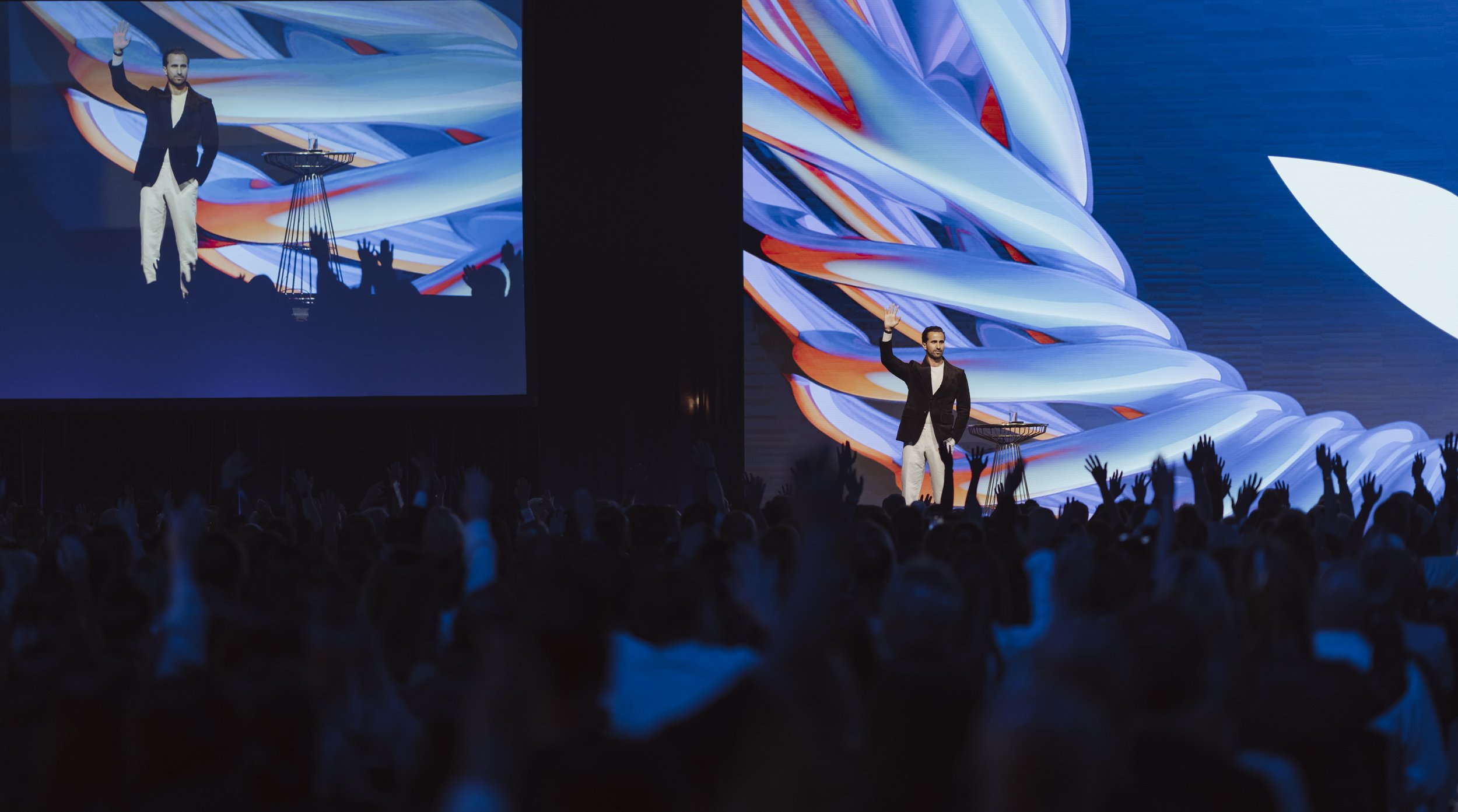 A man dressed in a black blazer and white pants waves on stage at a conference, with a large audience in front and a digital screen behind him showing abstract artwork.