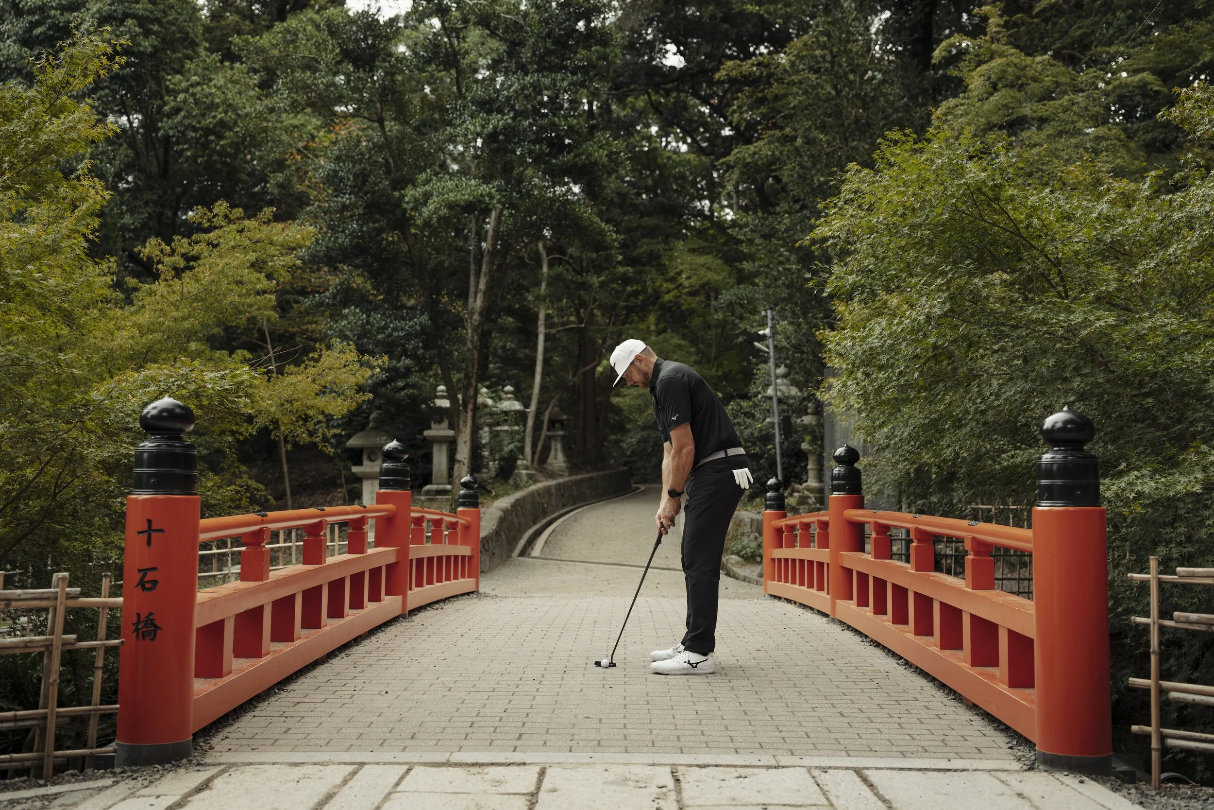 A man in golf attire is preparing to putt on a small bridge with orange railings over a pathway in a park, surrounded by green trees.