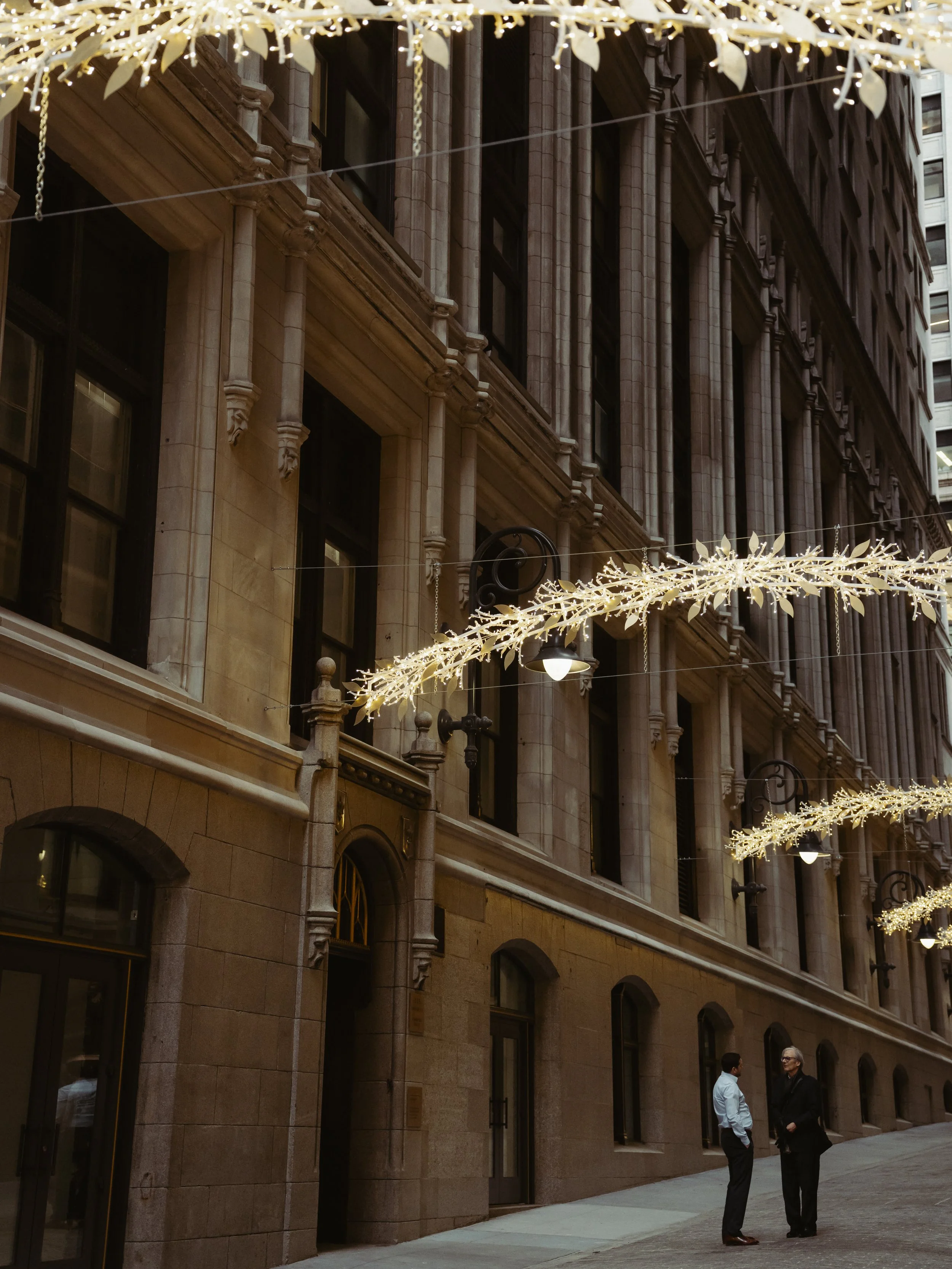 Two men in conversation on a city sidewalk decorated with hanging string lights and holiday lights in an urban setting.