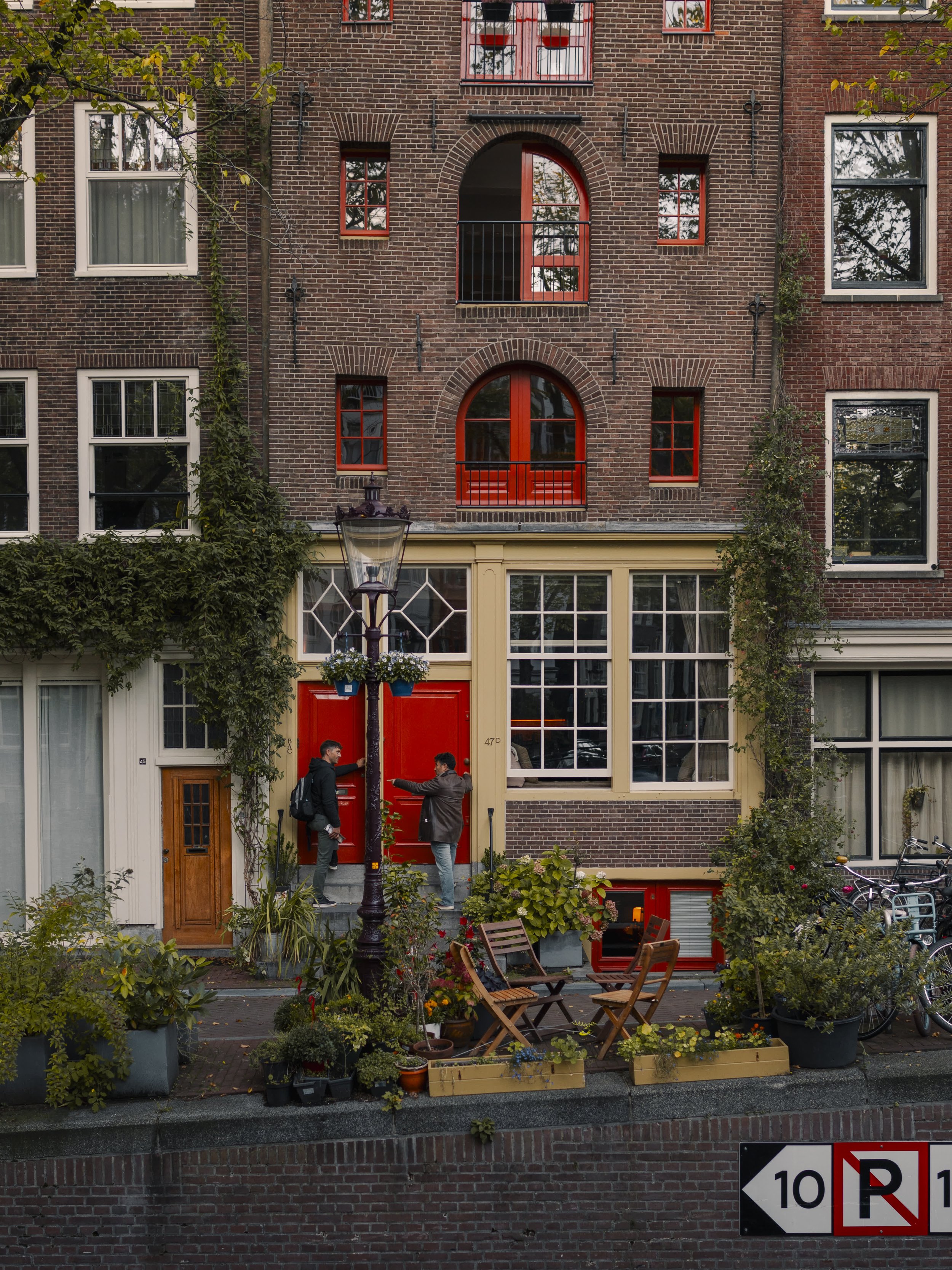 Two men talking in front of a red door on a building with large windows, surrounded by plants and outdoor furniture, with bicycles parked nearby.