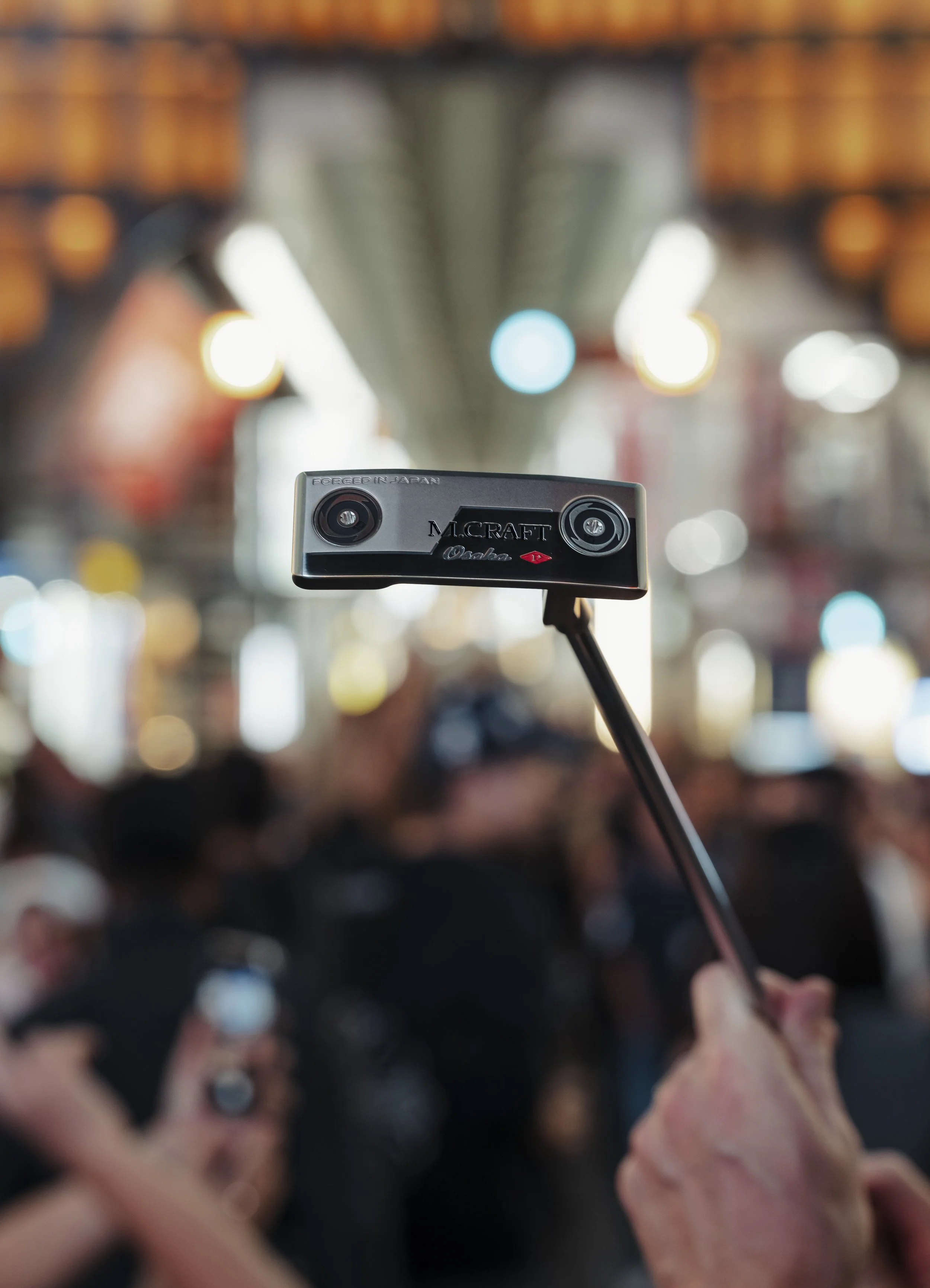 Close-up of a golf putter held up in a crowded indoor space with blurry audience and bright lights in the background.