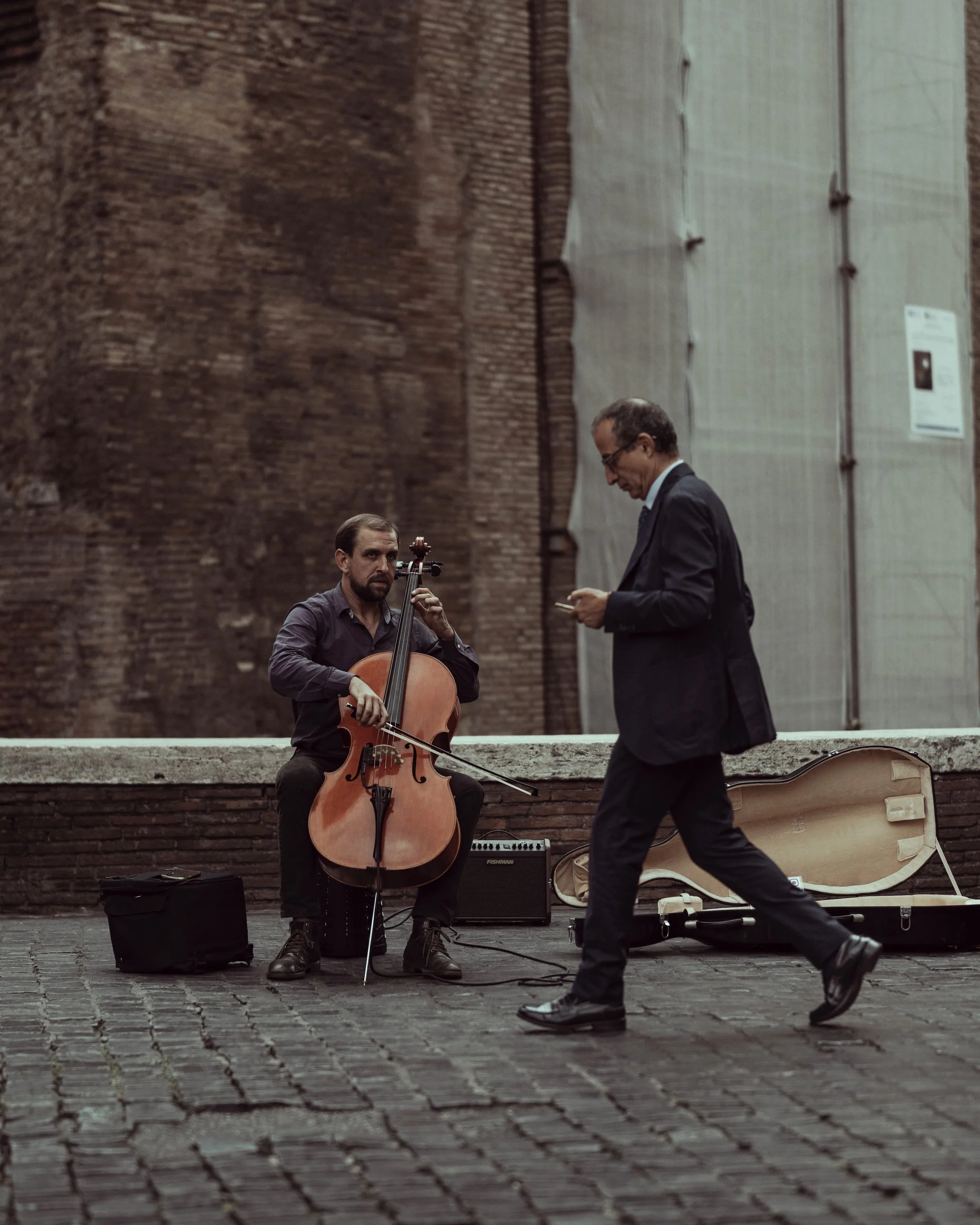 A street musician playing a cello while a man in a suit walking past him uses a smartphone, with a cello case open on the ground nearby.