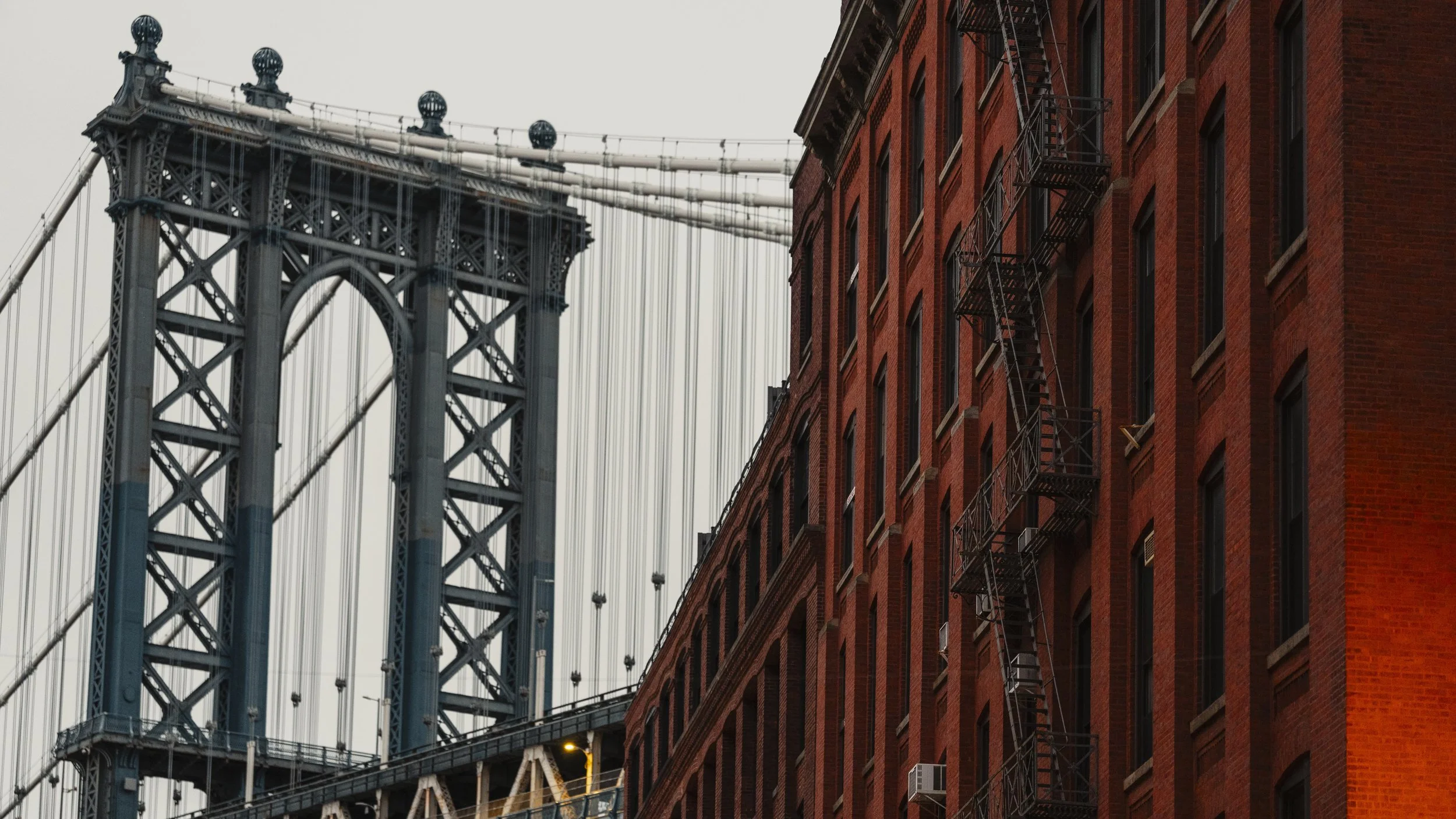 Partial view of a bridge with a tall, gray steel tower and suspension cables on the left, and a red brick building with fire escape stairs on the right.