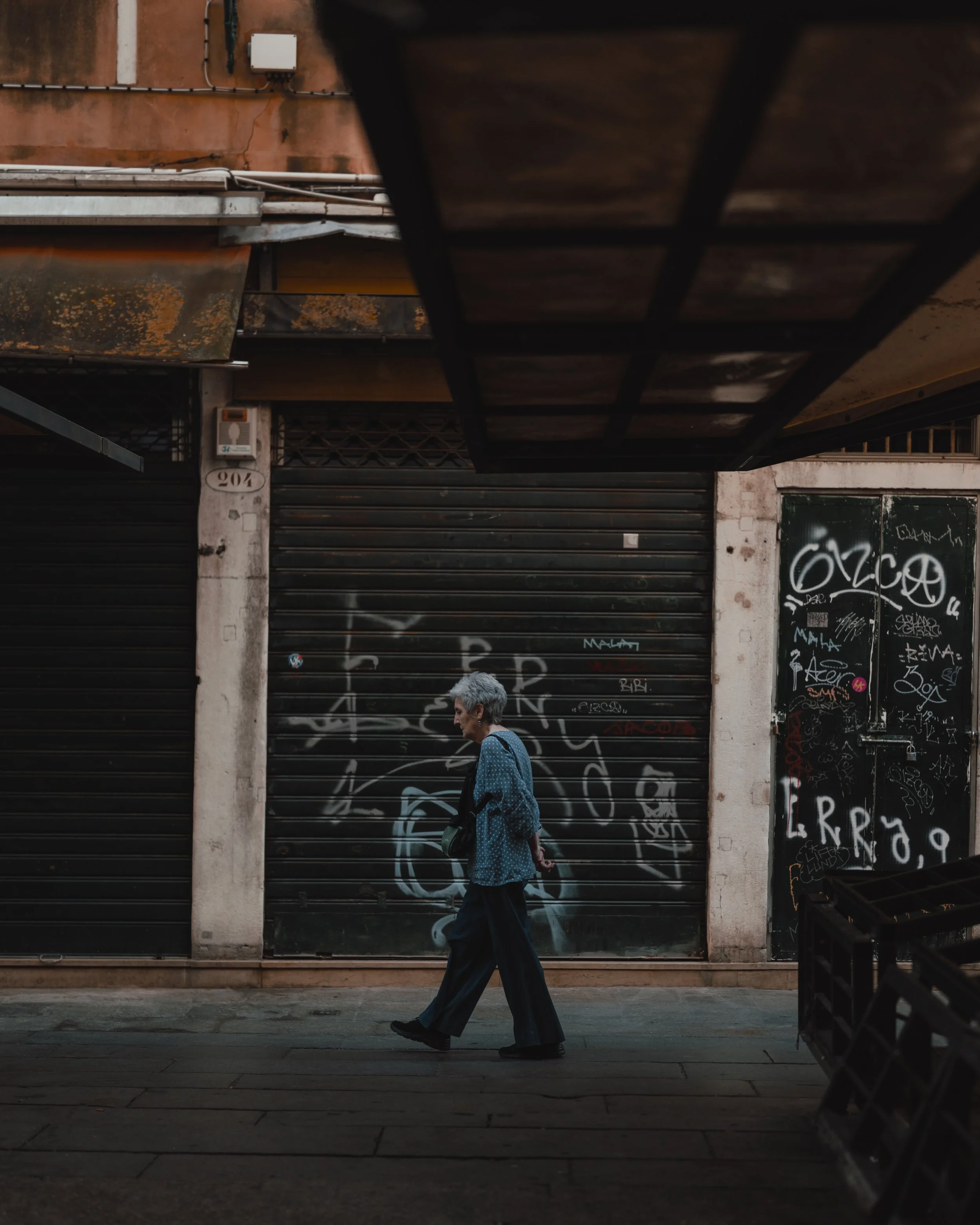An elderly woman walking past graffiti-covered metal shutters on a city street, with a dark, urban atmosphere.