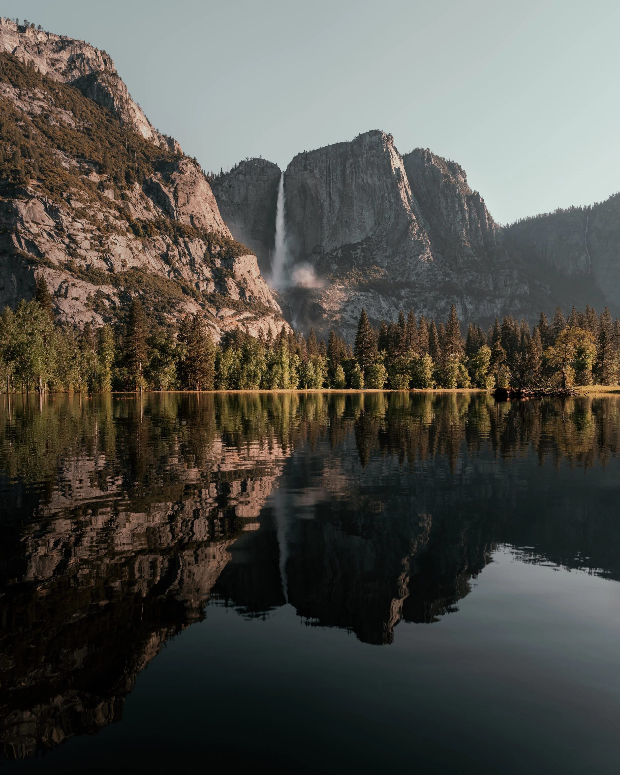 Photo of a serene lake with tall mountain cliffs and a waterfall in the background, surrounded by dense trees.