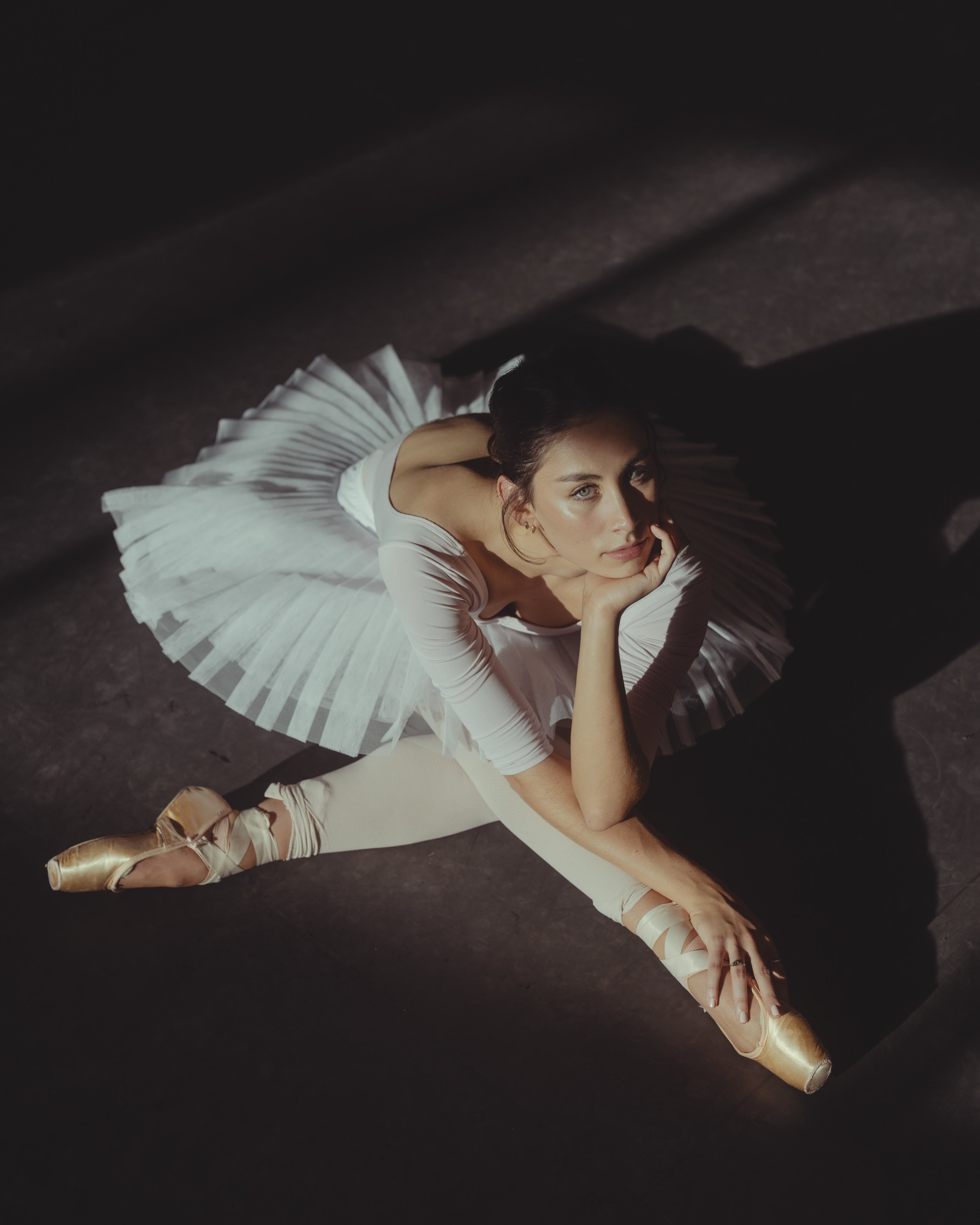 Ballerina in a white tutu and pointe shoes performing a split on the dark stage, resting her chin on her hand, top view shot.