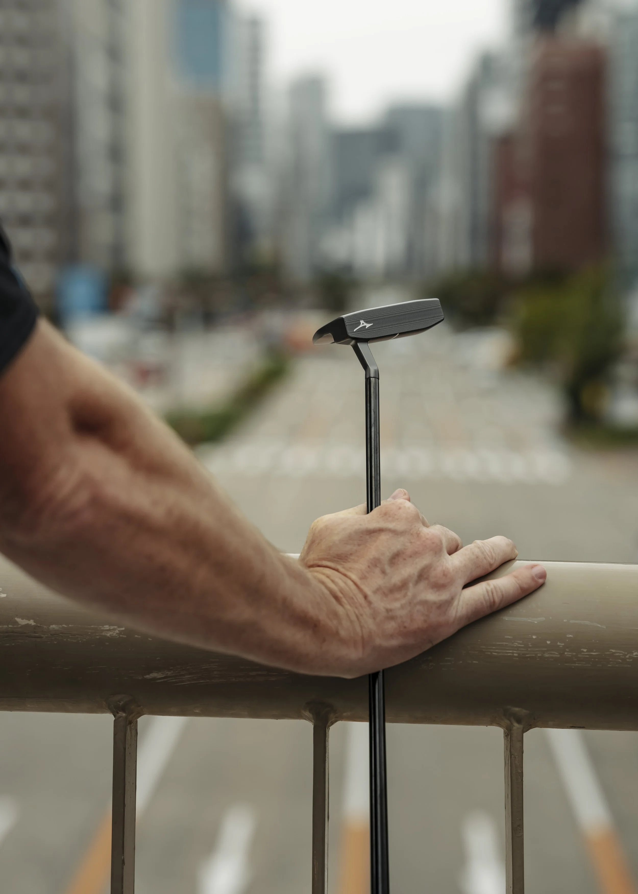Close-up of a person's hand holding a golf putter on a railing with a blurred city street background.