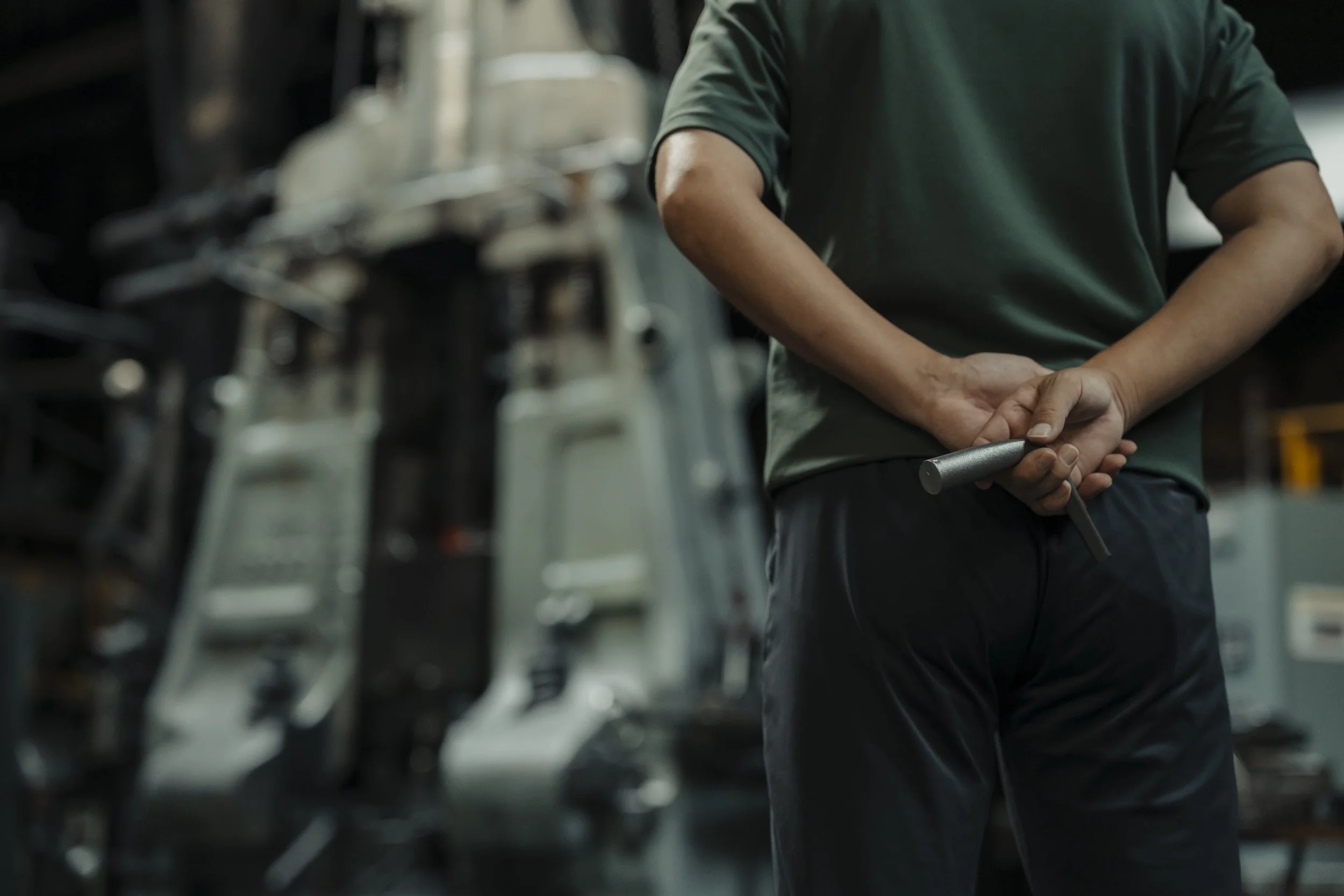 Person standing in a workshop or industrial setting, holding a wrench behind their back, with machinery and tools around.