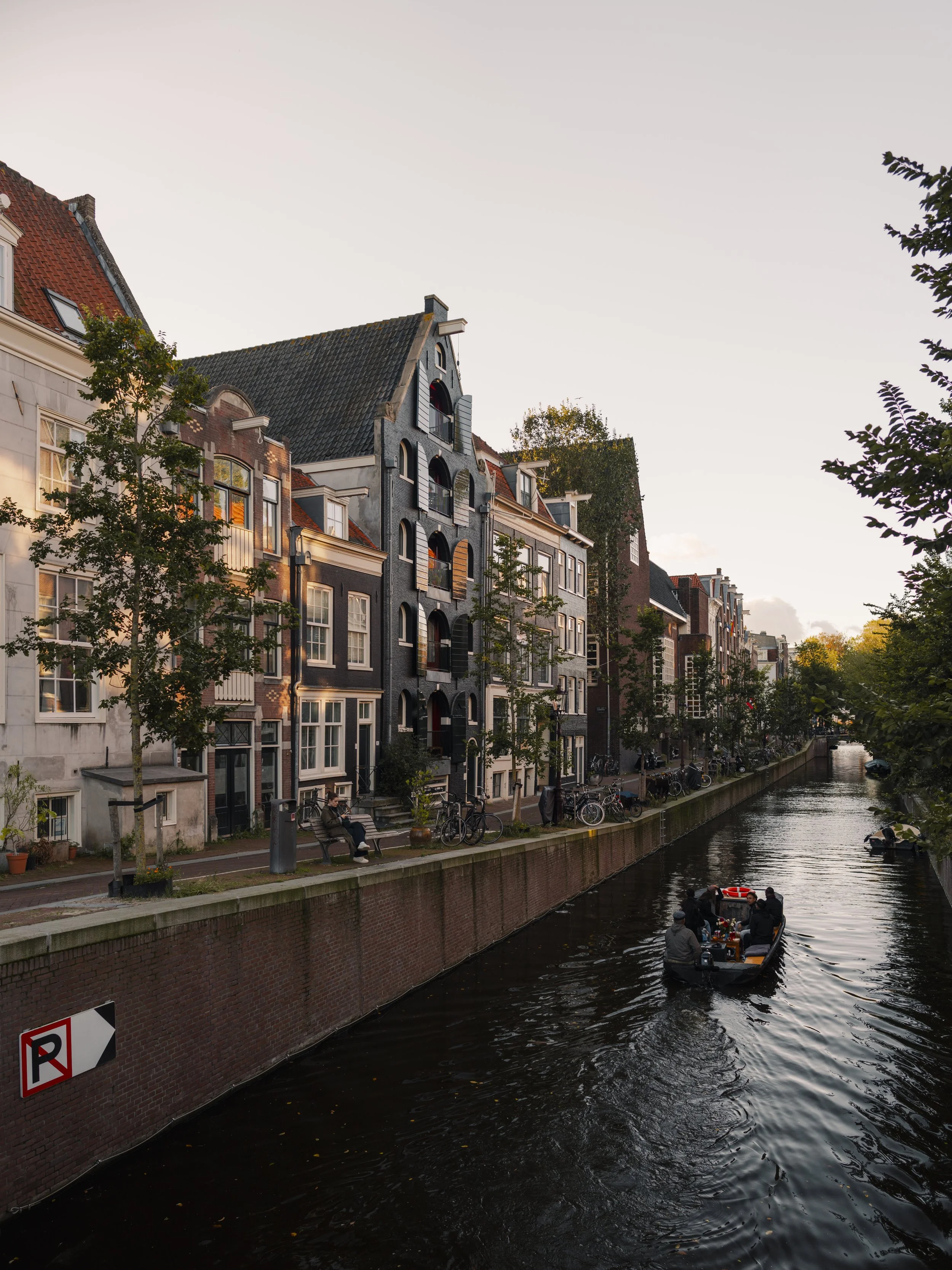 A canal lined with colorful old buildings, some with eliminated roofs, trees, a boat with people, and a person sitting on a bench along the sidewalk at sunset in a European city.
