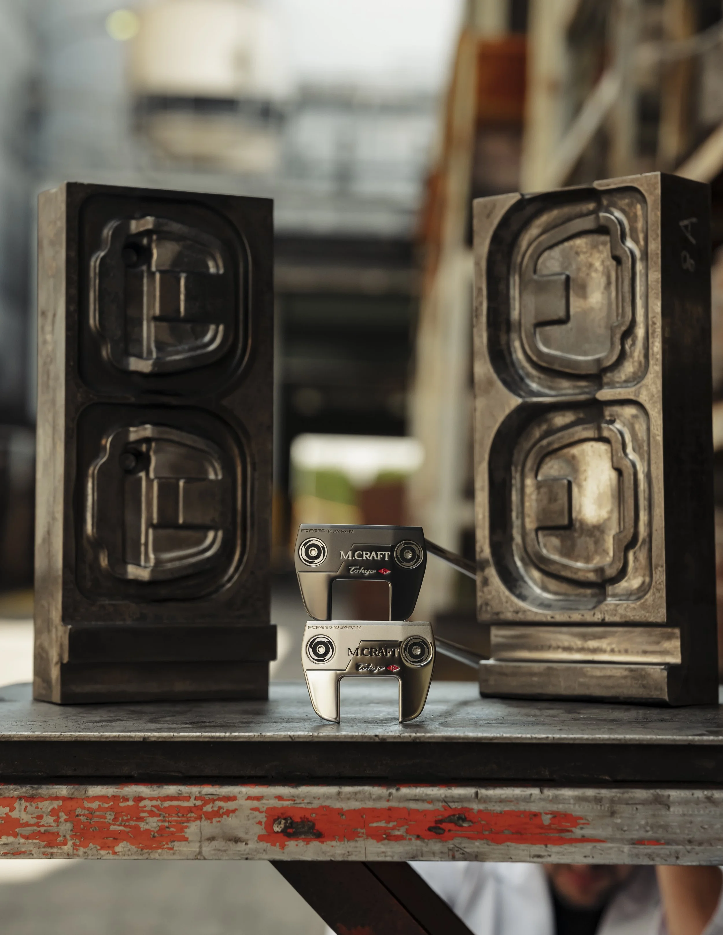 Two metallic golf putters with black and silver heads positioned in front of two molds for golf club heads on a work table.