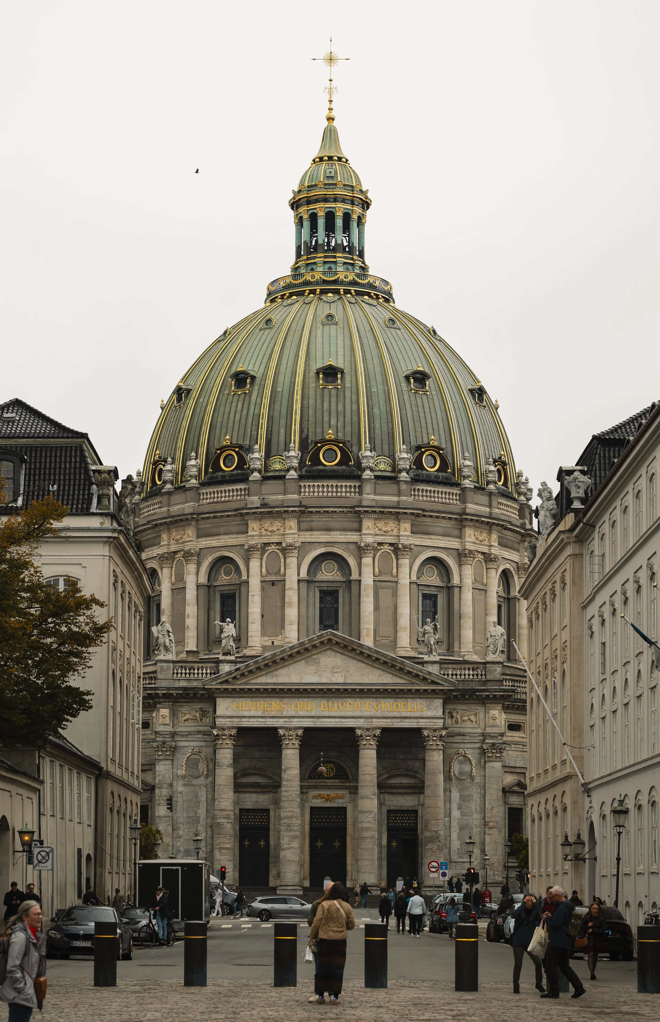Front view of a historic church building with a large, greenish and gold dome, classical columns, and statues on top, surrounded by city street with pedestrians and parked cars.