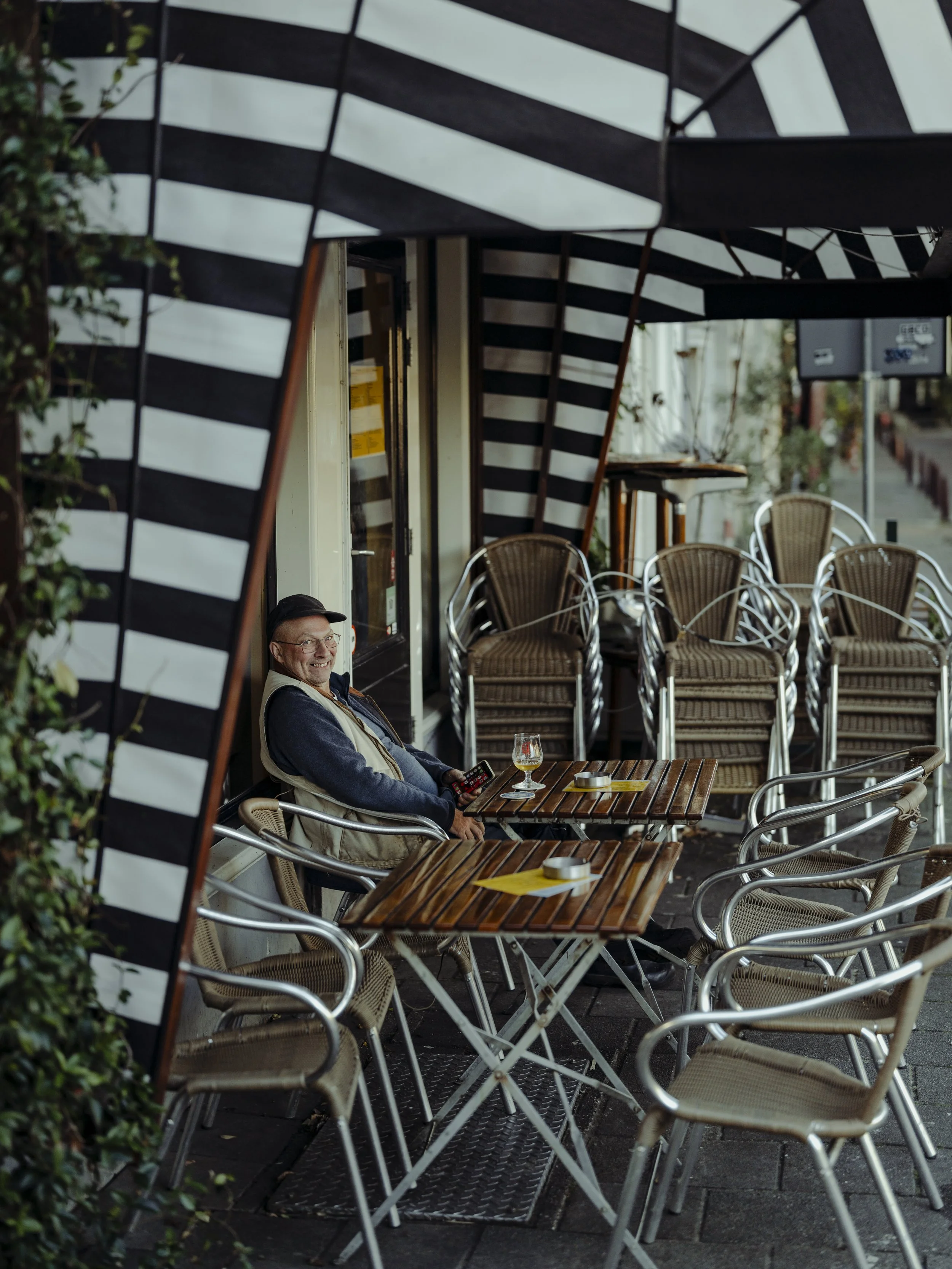 A man sitting at an outdoor cafe table, smiling, holding a remote control, with drinks and yellow napkins on the table, under a striped black and white patio umbrella, with stacked chairs nearby.