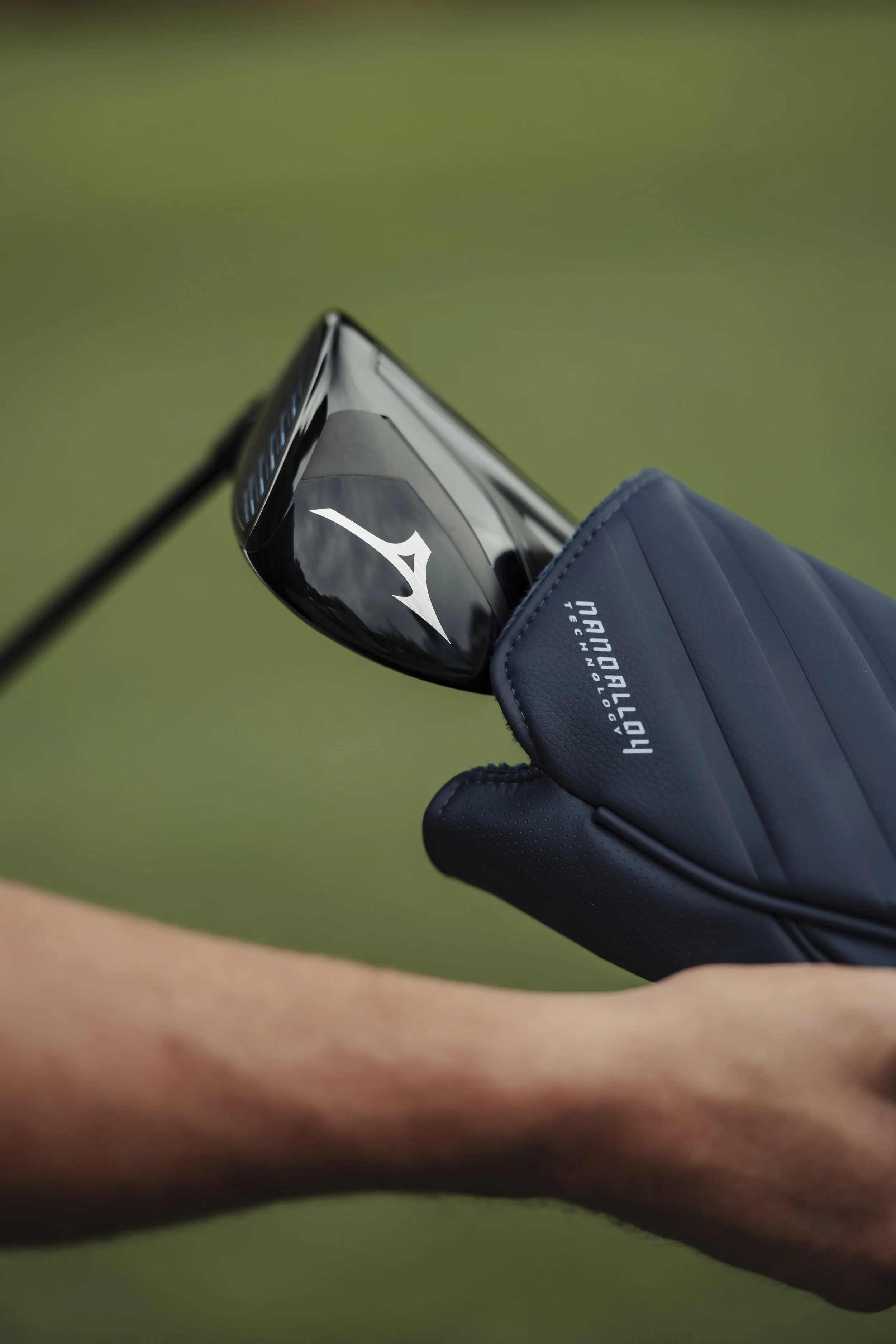 Close-up of a golf club in a headcover with a hand holding it, on a golf course with green grass in the background.