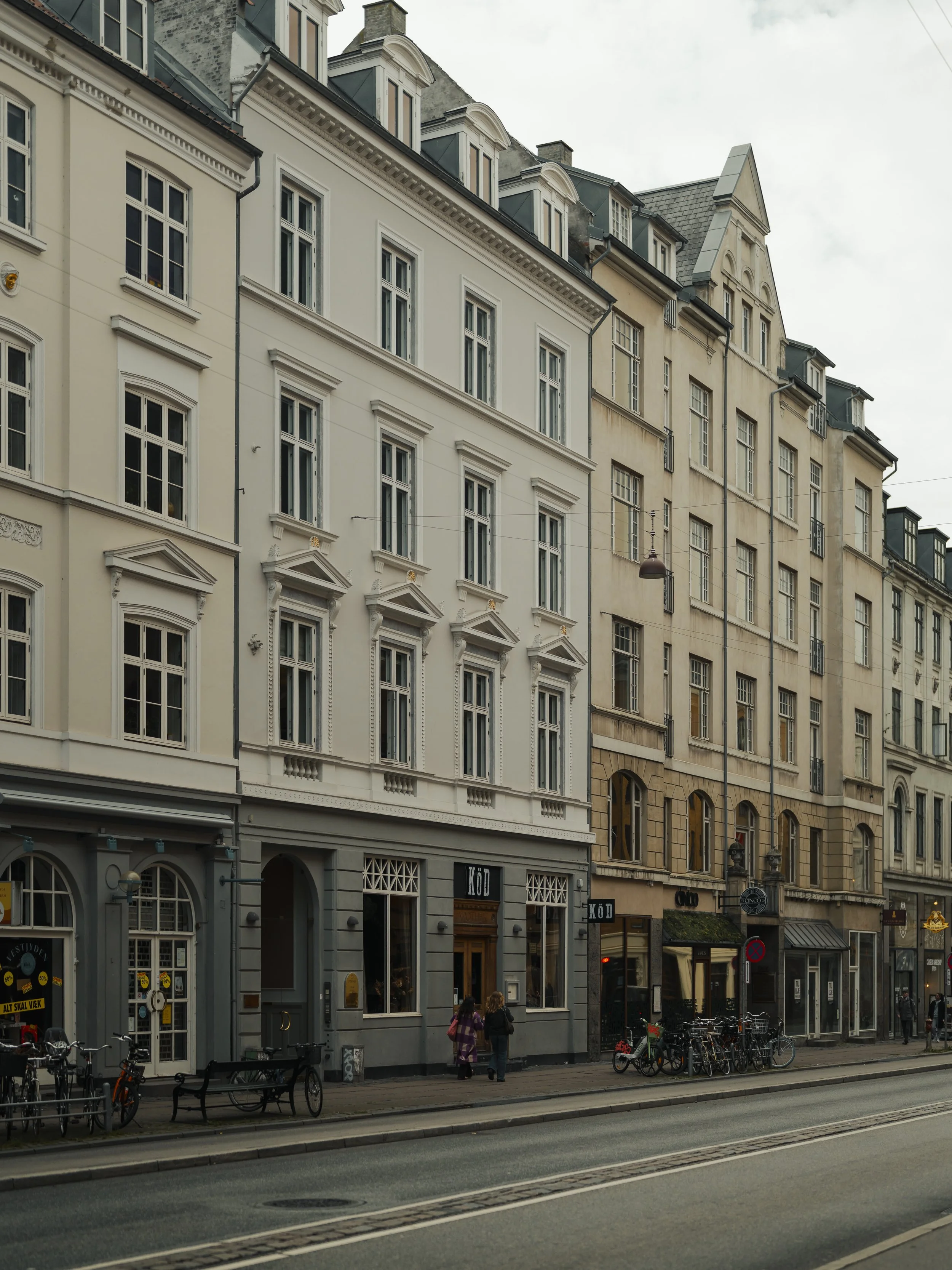 European city street with multi-story historic buildings, bicycles parked along the sidewalk, and pedestrians walking.