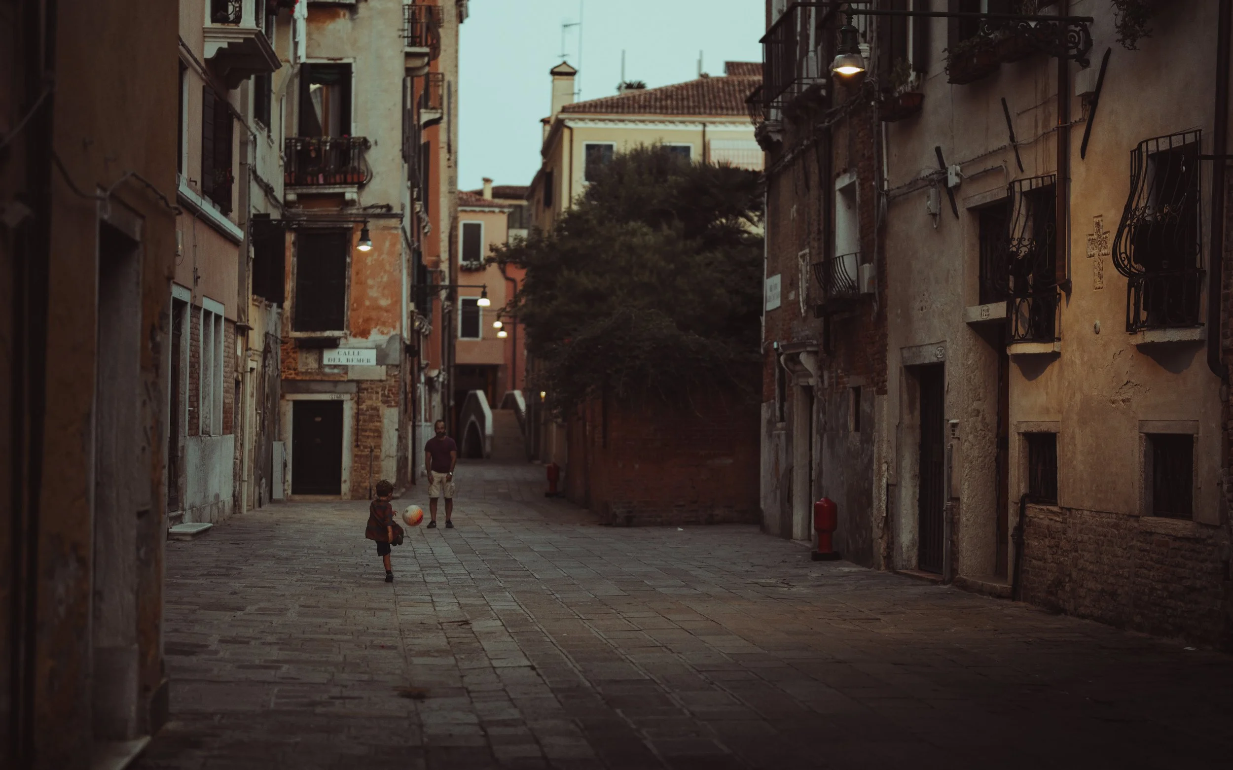 A narrow European alleyway with old, weathered buildings on both sides, lit by street lamps, with a man and a child playing with a ball in the distance.