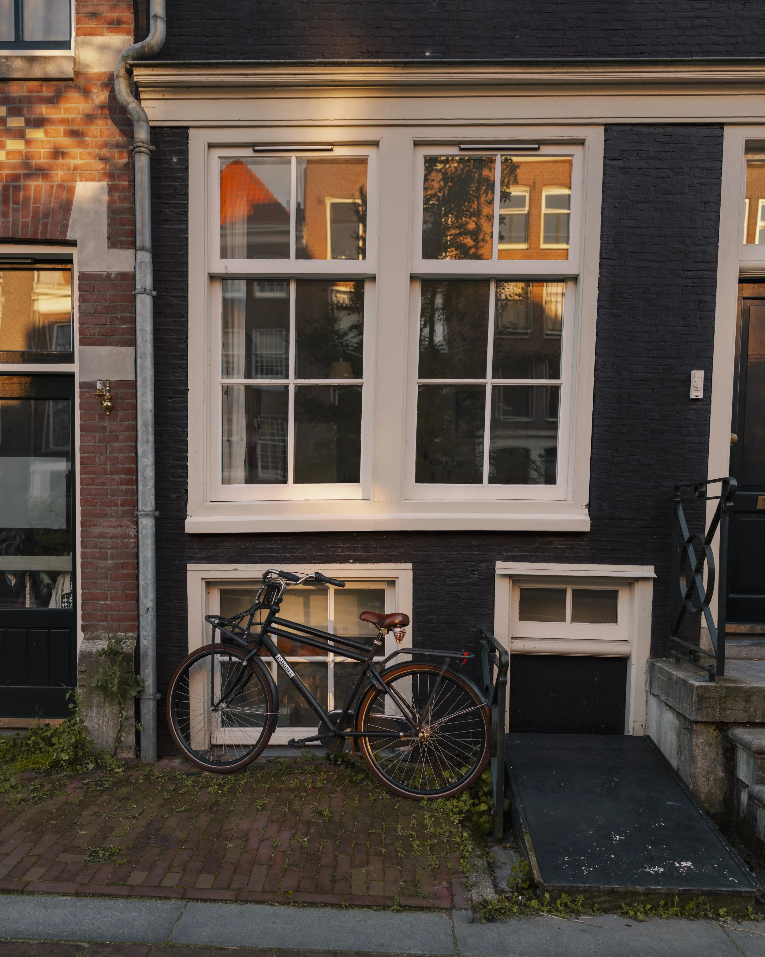 A black bicycle with a brown saddle and handlebars parked outside a brick and black-painted building with large white-framed windows, illuminated by warm sunlight.