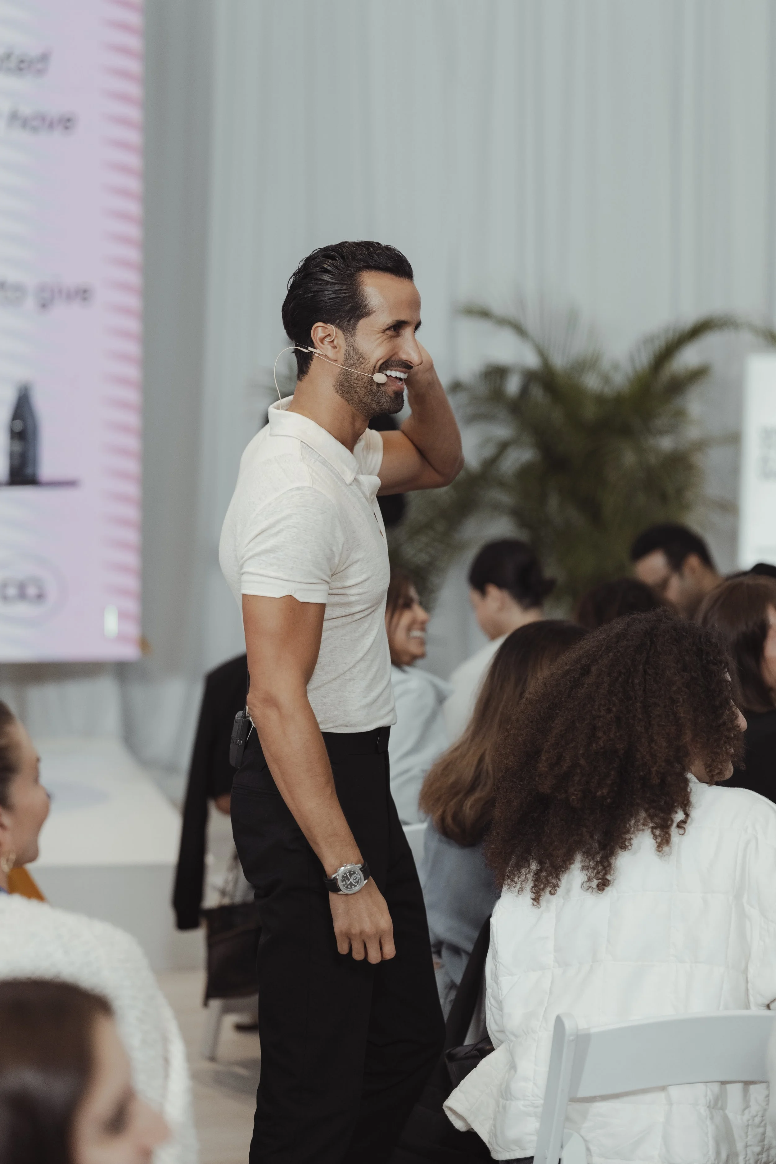 A man with dark hair and a beard wearing a white polo shirt and black pants is smiling and standing at a conference or seminar, using a headset microphone. Several other people are seated around him, listening and engaging in the event.