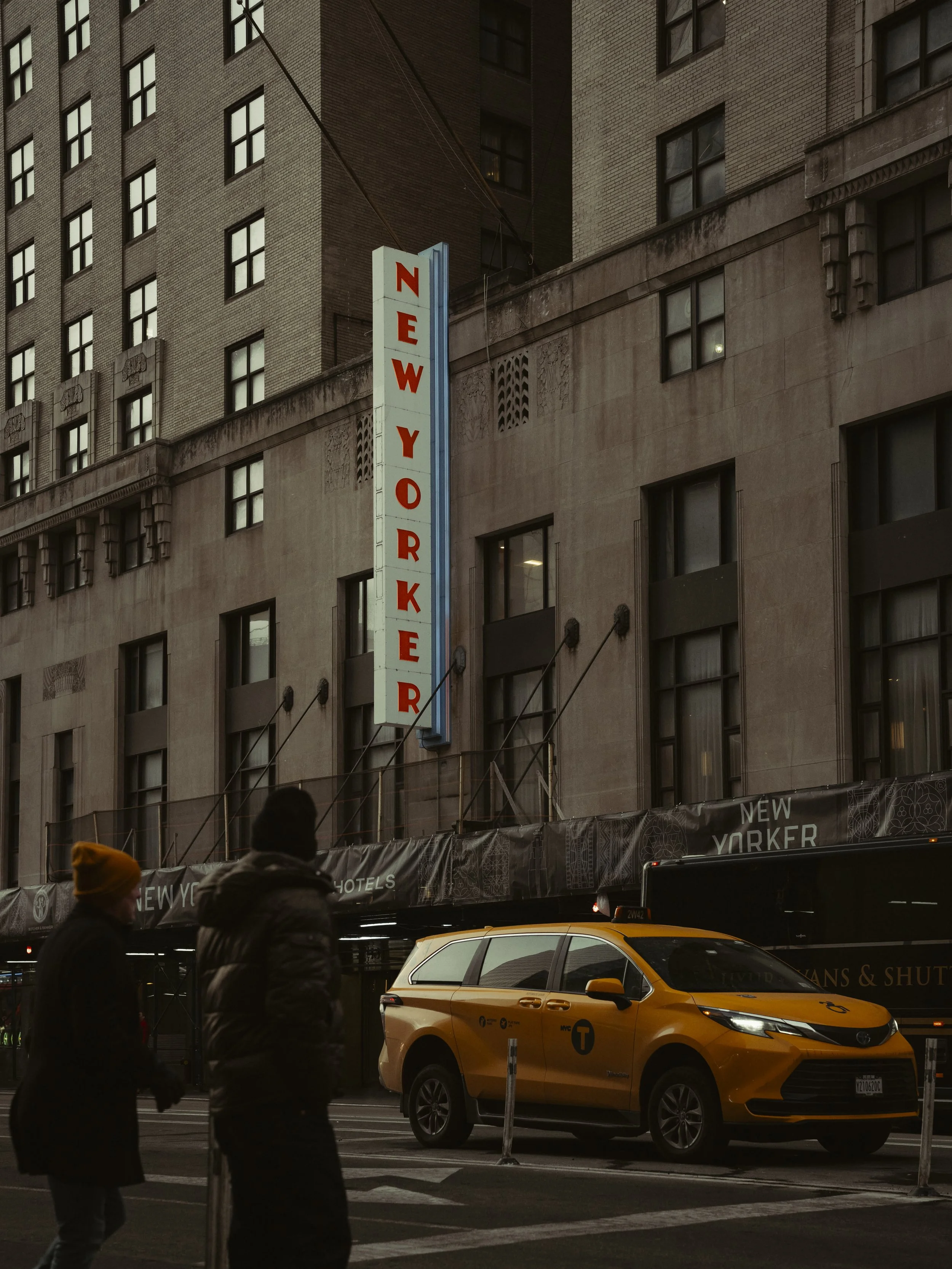 Street scene in New York City showing a building with a vertical sign reading "New Yorker," two pedestrians walking, and a yellow NYC taxi in front.