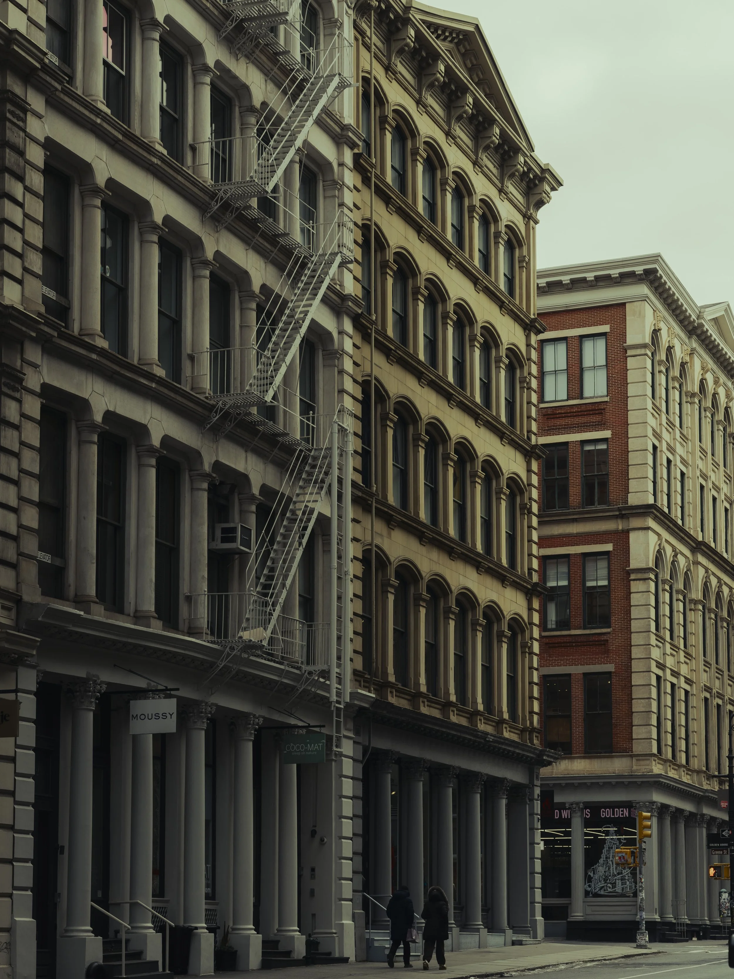 Street view of historic buildings with fire escapes on a city sidewalk, two people walking, storefronts at the ground level.