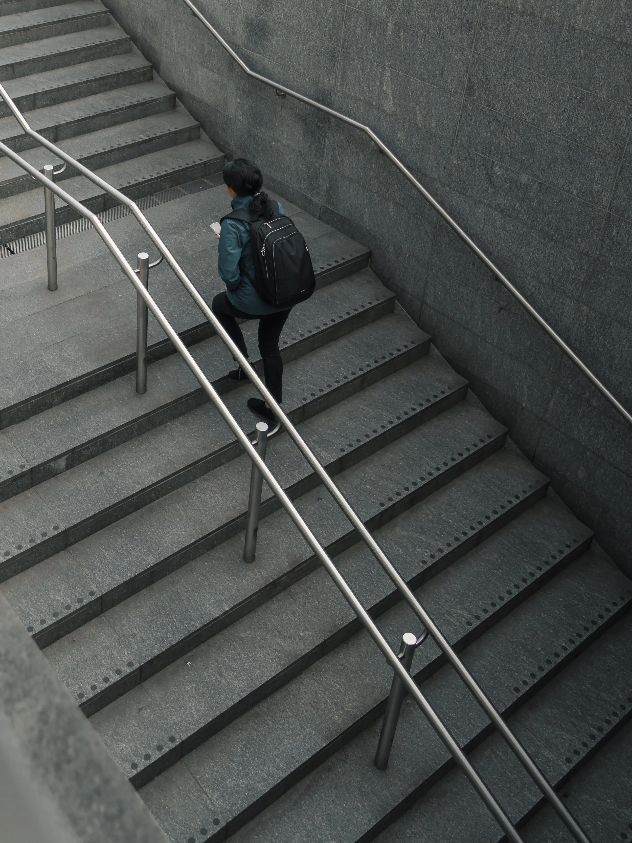 A woman with a black backpack descending a gray stone staircase with metal handrails in an urban setting.