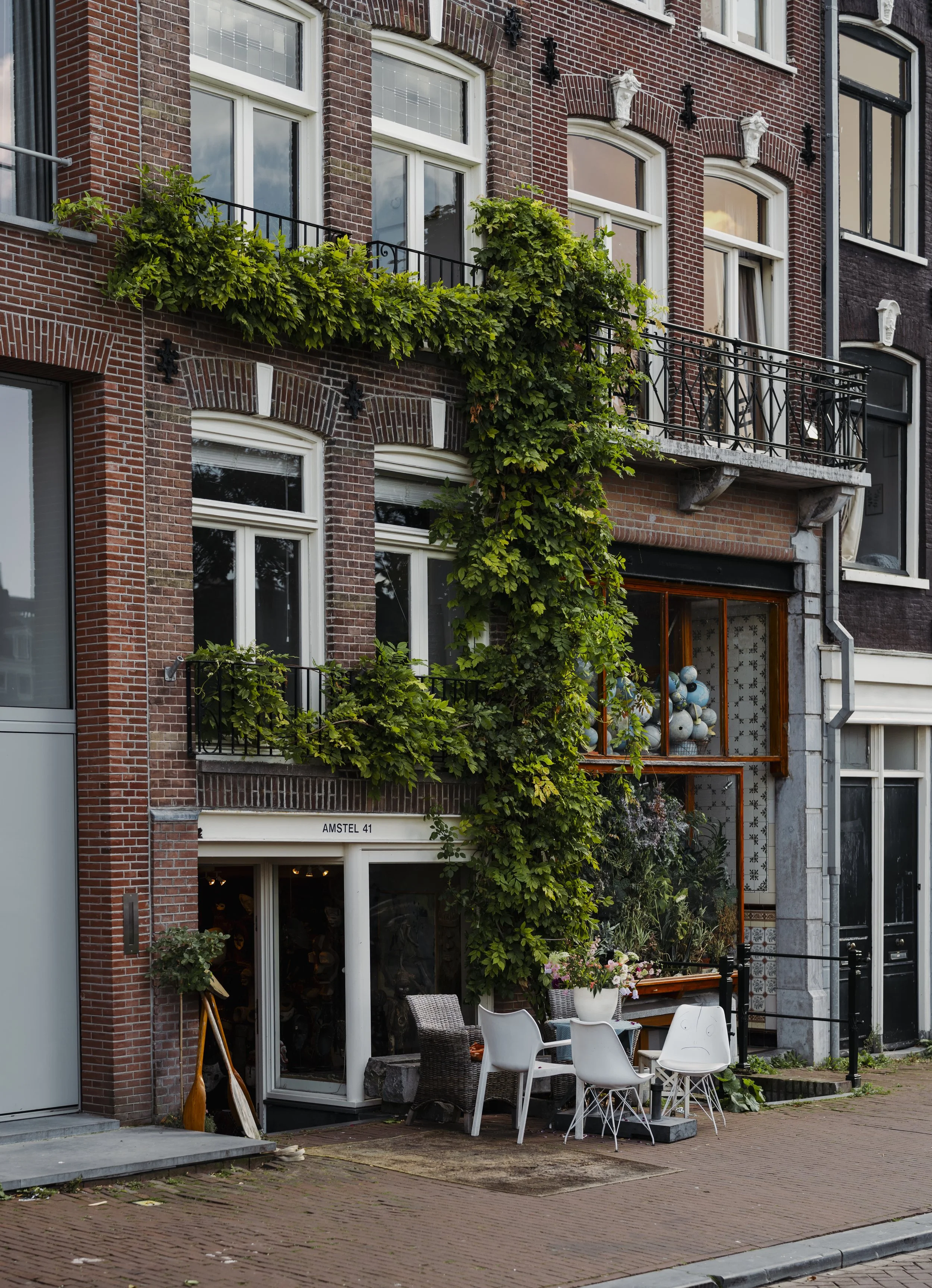 A brick building with green plants growing on the balconies and around the windows. Small outdoor seating with chairs and a table in front of the building. Display of vases and plants inside a large window, with the address 'Amstel 41' on the buildin