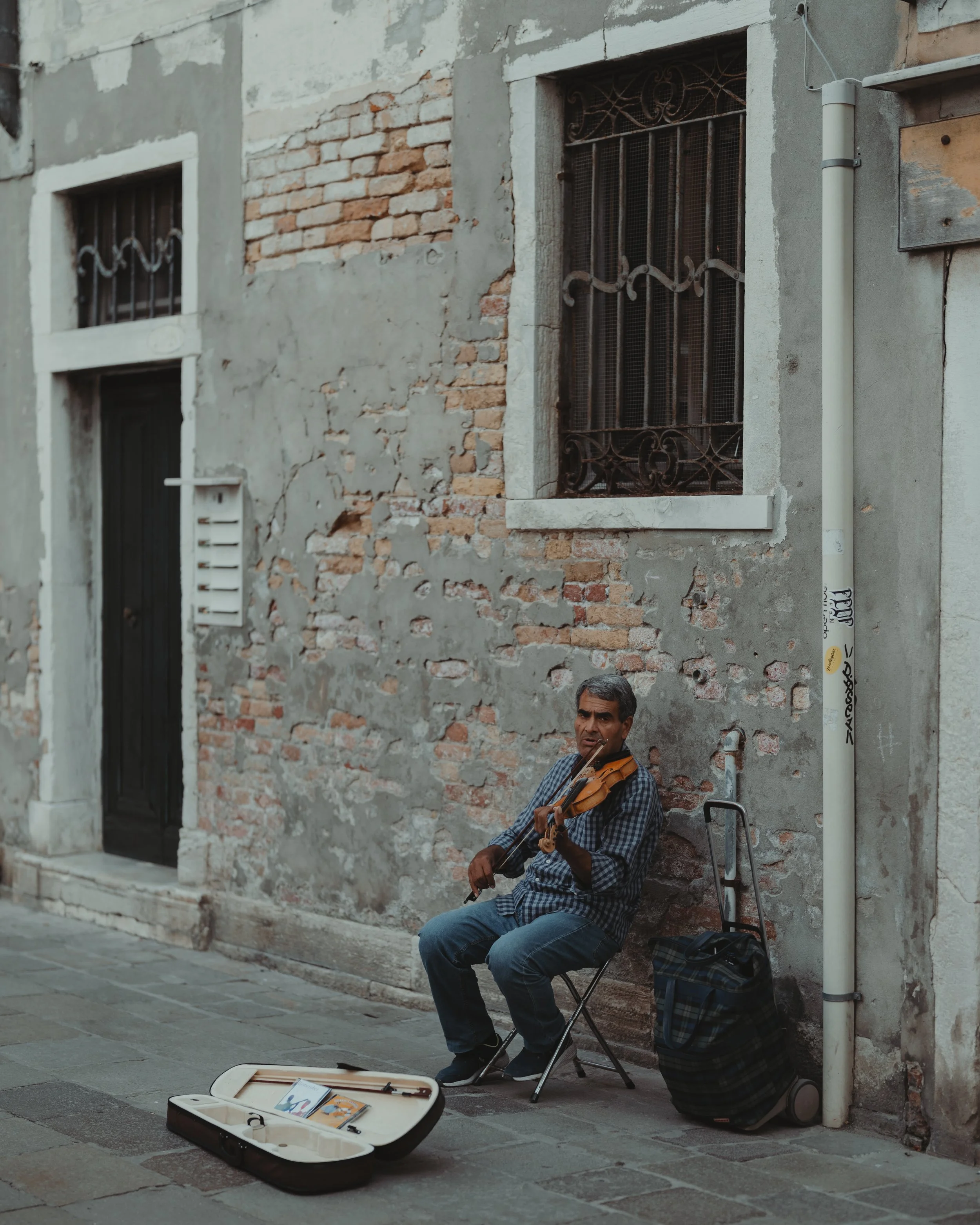 A man playing violin while sitting on a small foldable chair against a weathered brick and concrete wall on a narrow street.