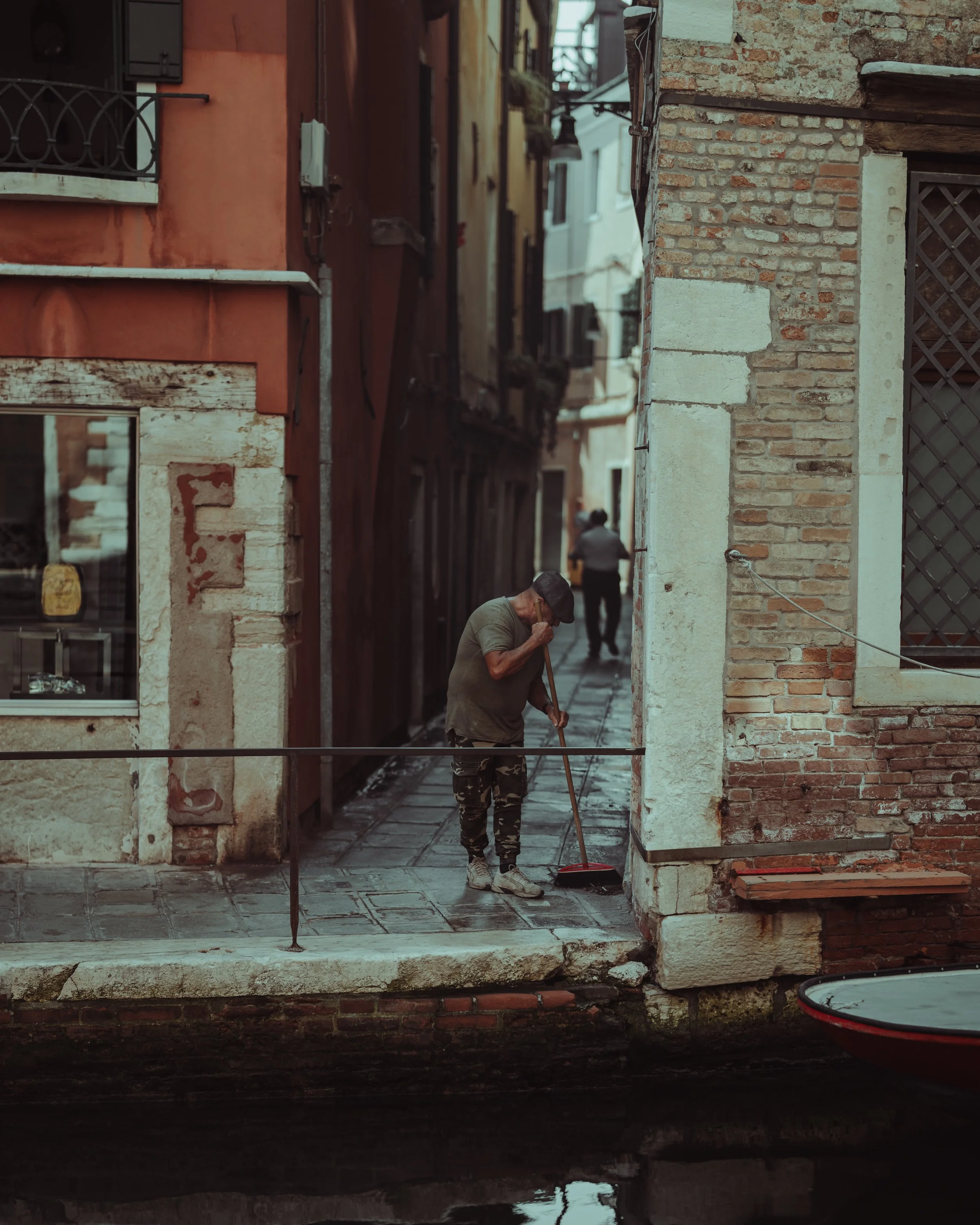 A man is sweeping the sidewalk next to a canal in Venice, Italy, with colorful buildings and narrow alleyways in the background.