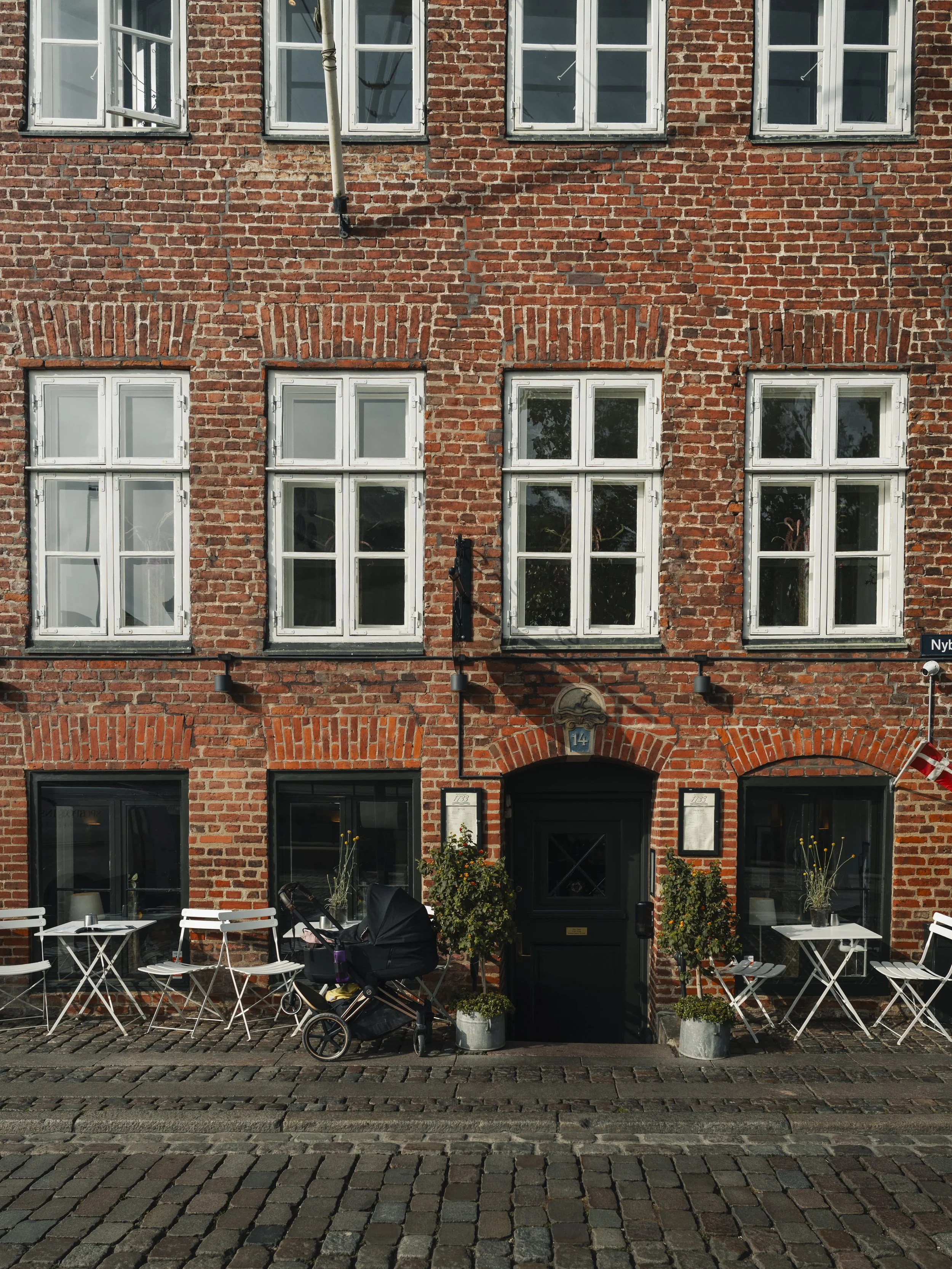 Red brick building with white-framed windows, outdoor tables and chairs, potted plants, and a stroller parked outside.